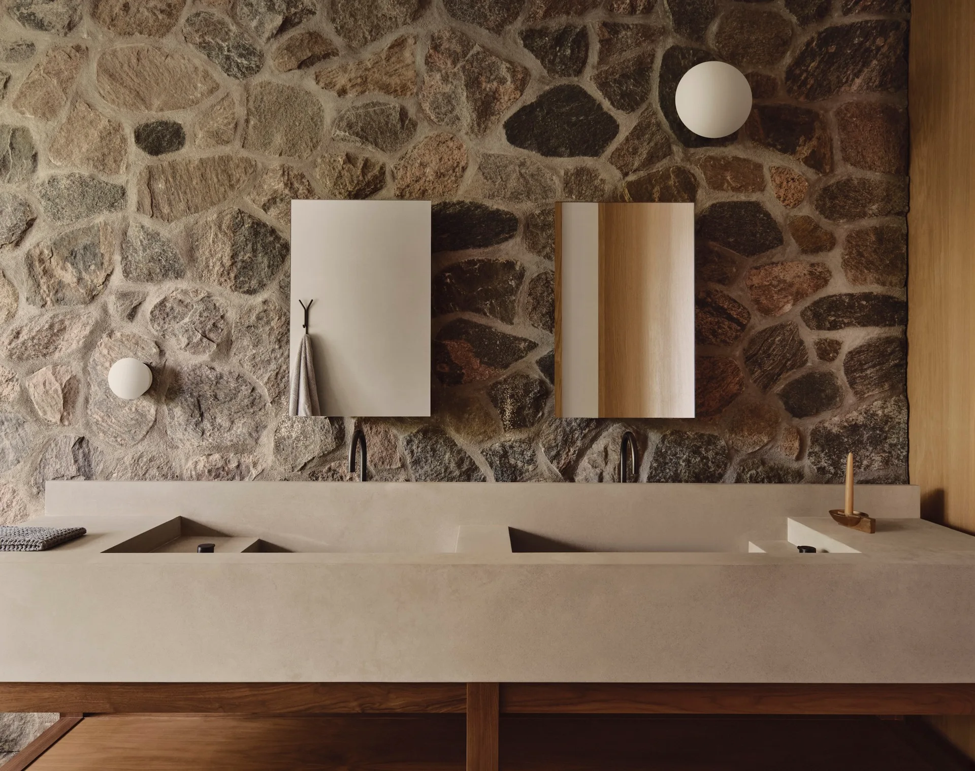 A bathroom with a long beige concrete double vanity featuring integrated sinks against a wall clad in rustic, irregular natural stone.