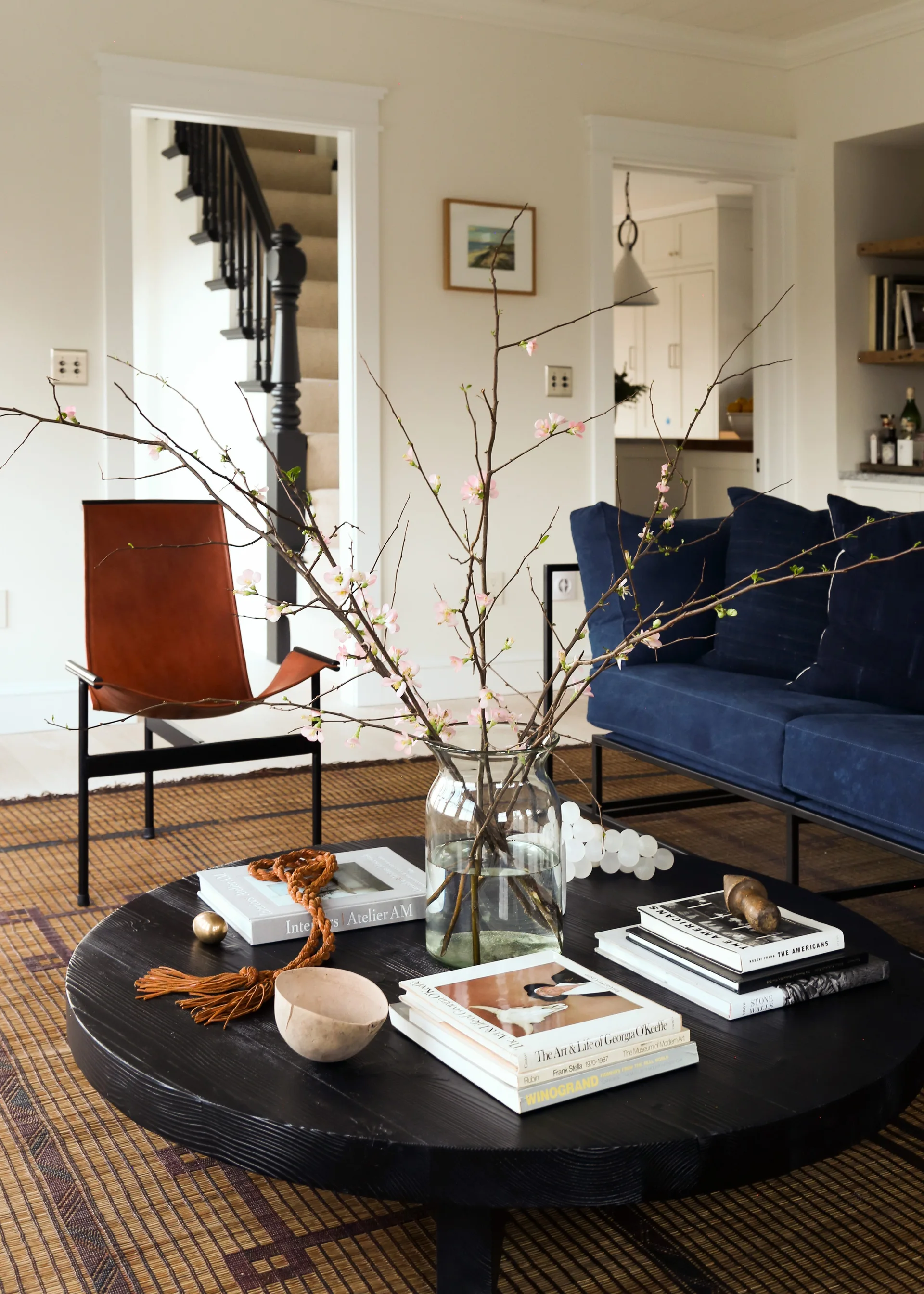 A living room featuring a black round coffee table topped with books and a vase of pink blossoms, a navy blue sofa, and a tan leather accent chair.