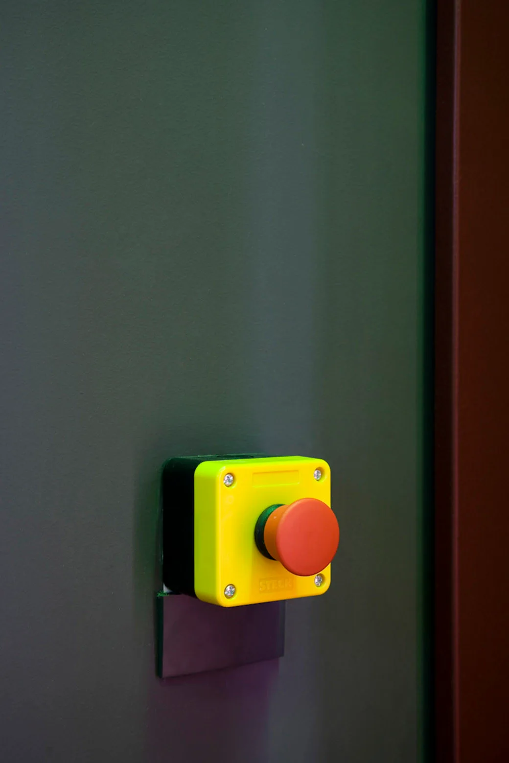 A bright yellow and red industrial emergency stop button is mounted on a smooth, matte dark green painted wall.