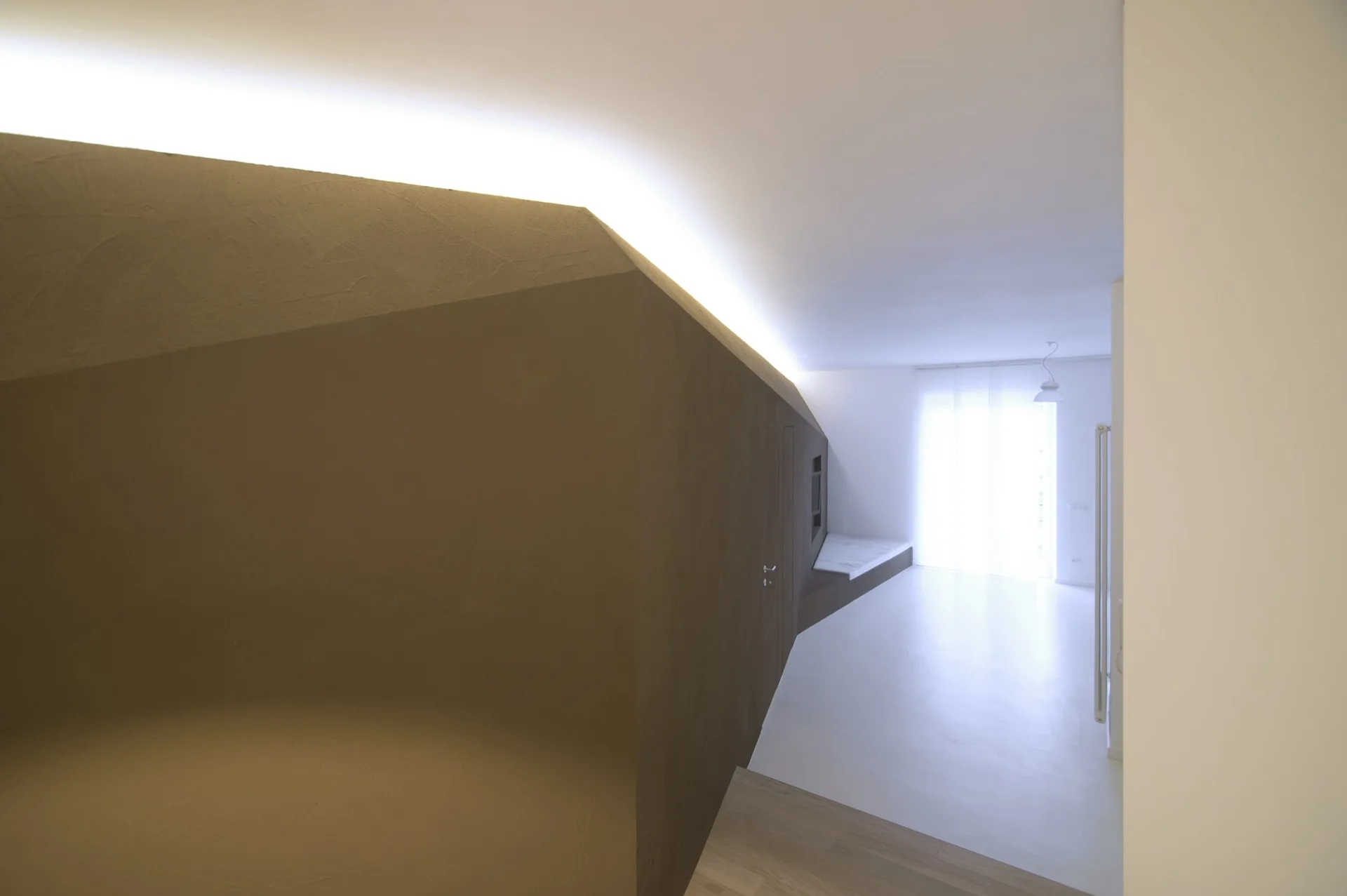A minimalist interior space featuring a large angled brown wall with bright cove lighting along the top leading toward a sunlit window.