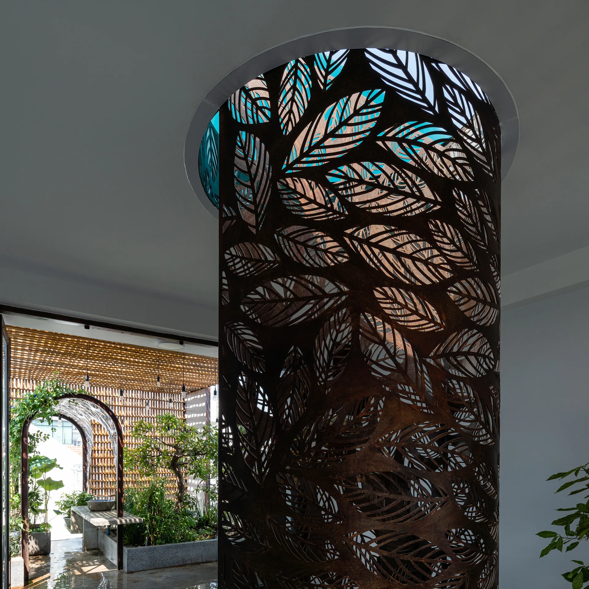 A cylindrical rusted metal screen with a laser-cut leaf pattern sits beneath a circular skylight, with a sunlit garden and wooden trellis visible behind.