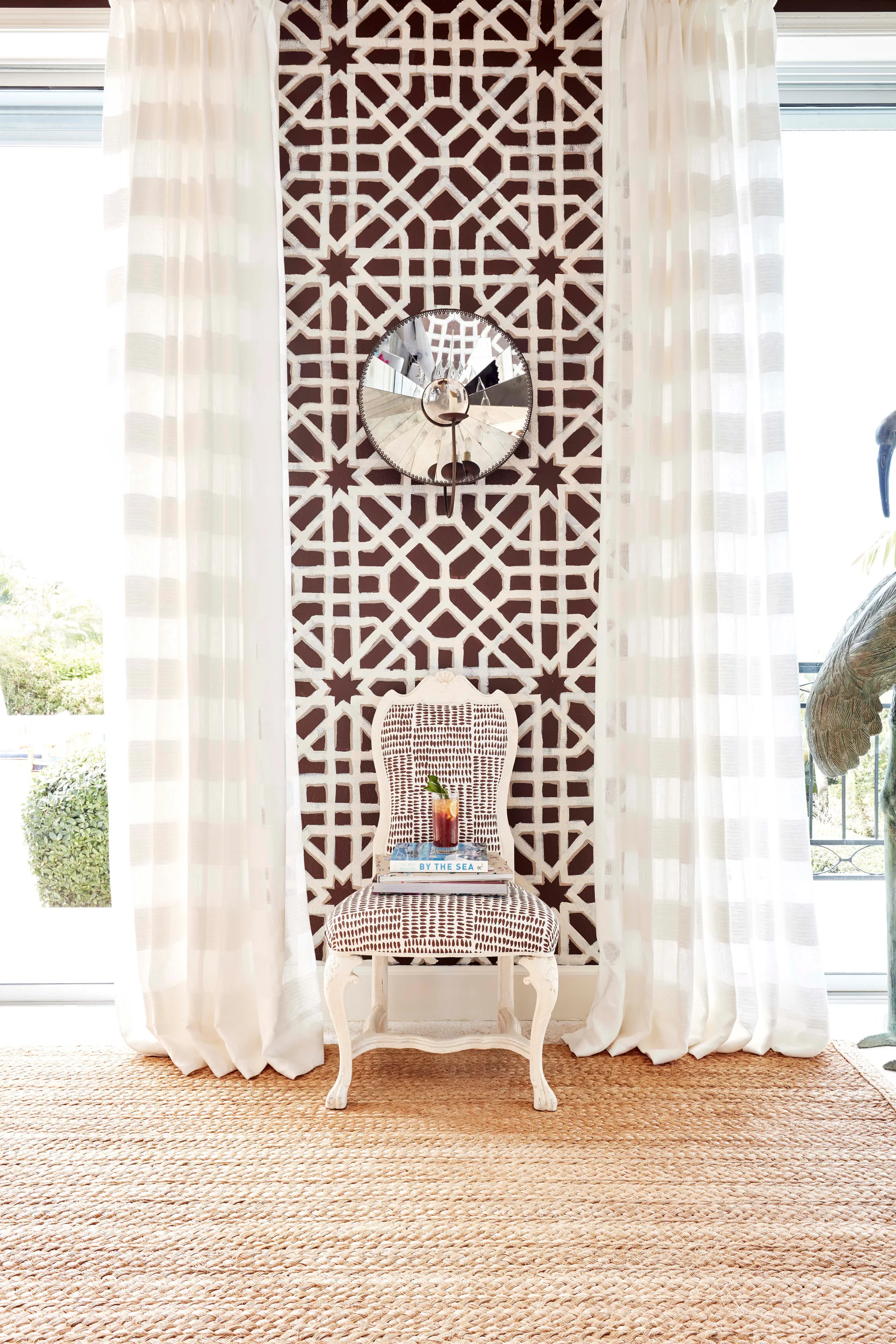 A patterned armchair sits in front of a brown geometric wallpapered wall strip flanked by white floor-length curtains on a woven jute rug.