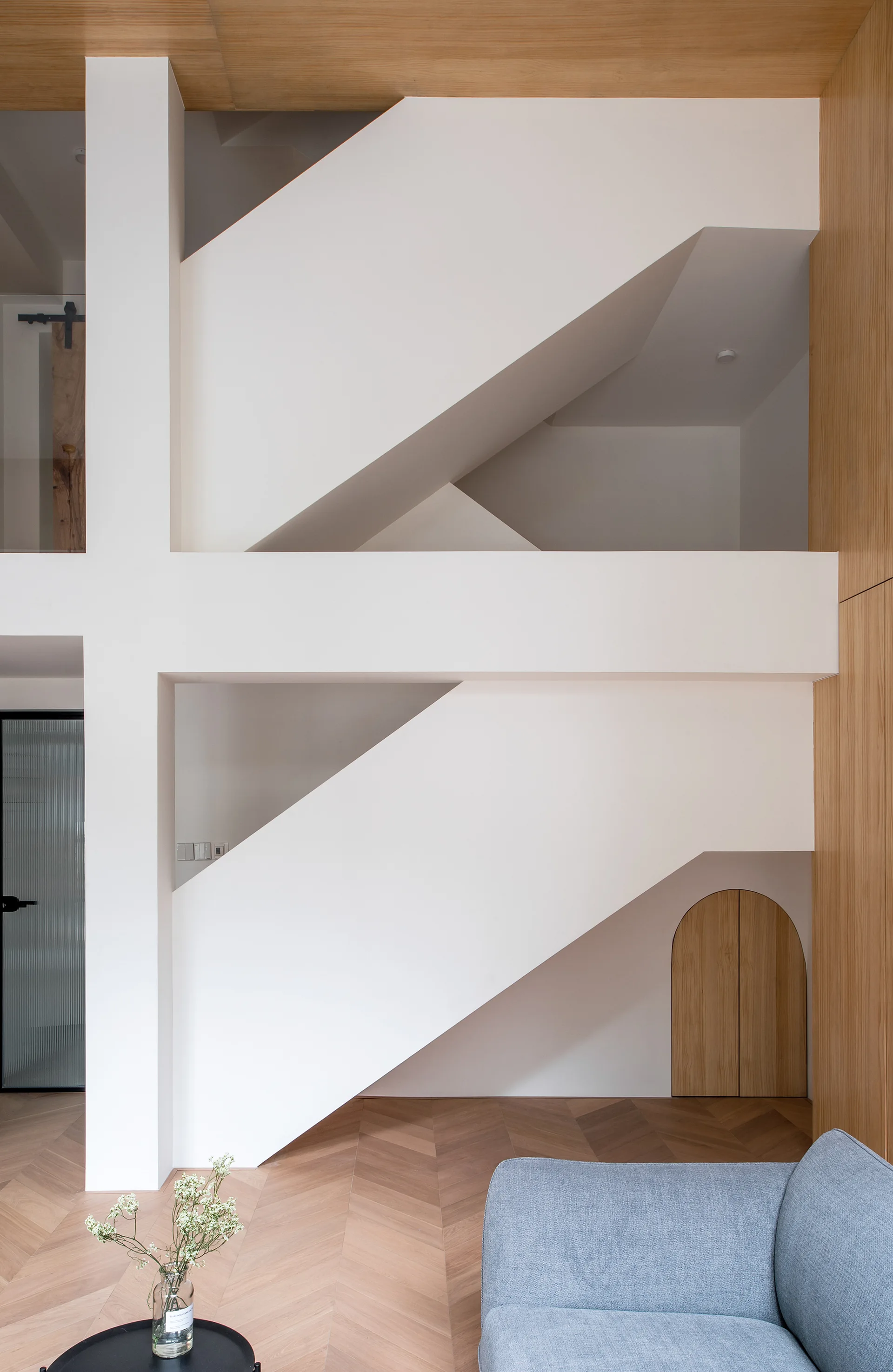 A modern white geometric staircase sits above herringbone wood floors next to a small arched wooden door built under the stairs.