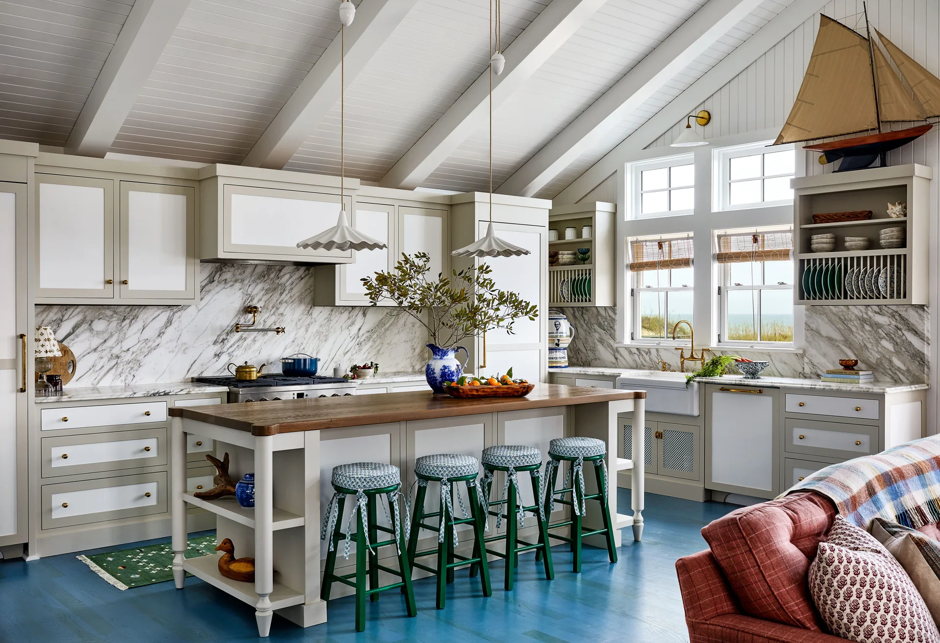 A coastal kitchen featuring blue stained wood floors, a wood-topped island with green stools, grey cabinetry, and a vaulted ceiling with beams.