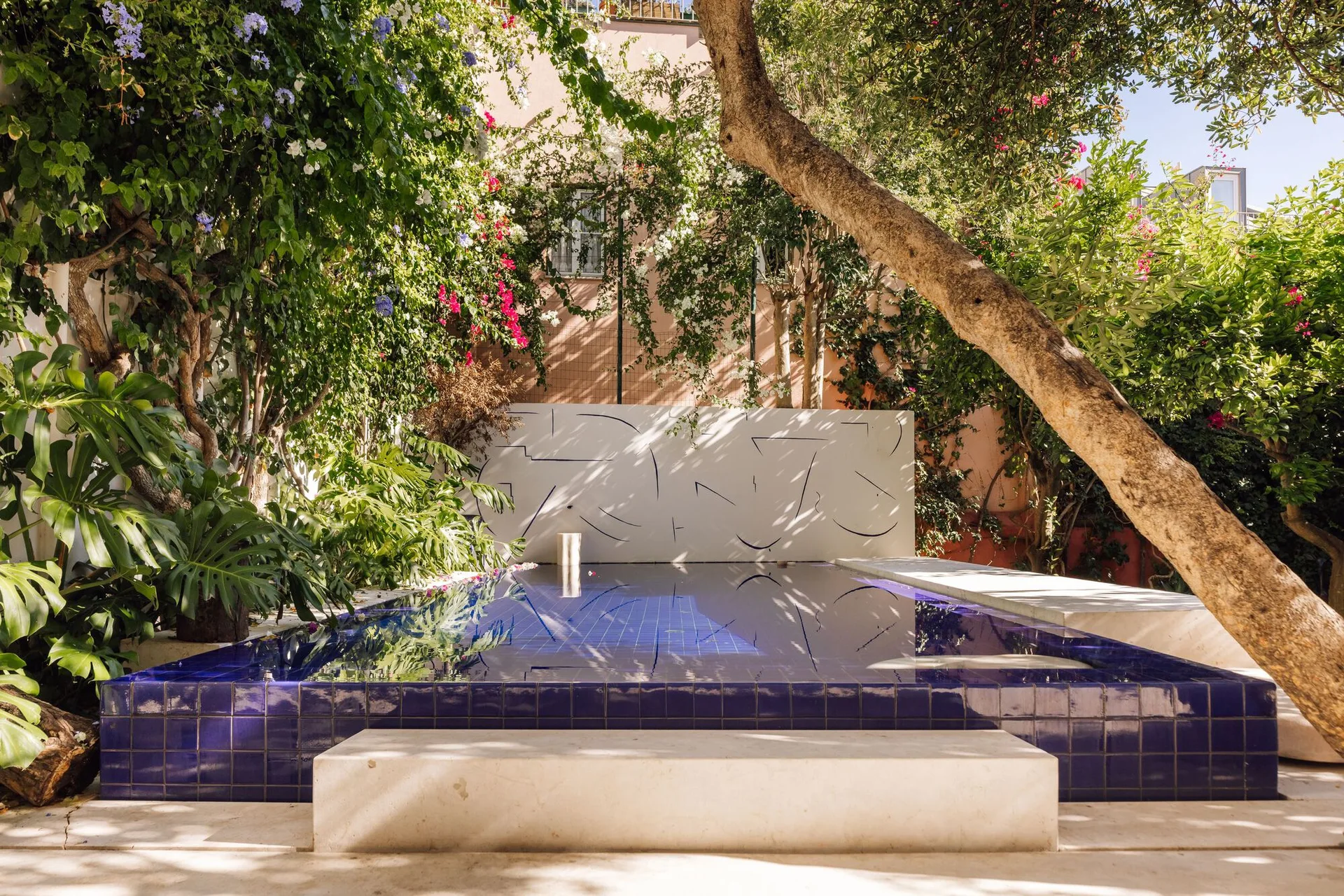 A rectangular swimming pool with deep blue tiles sits in a lush garden next to a white wall adorned with black minimalist line art.