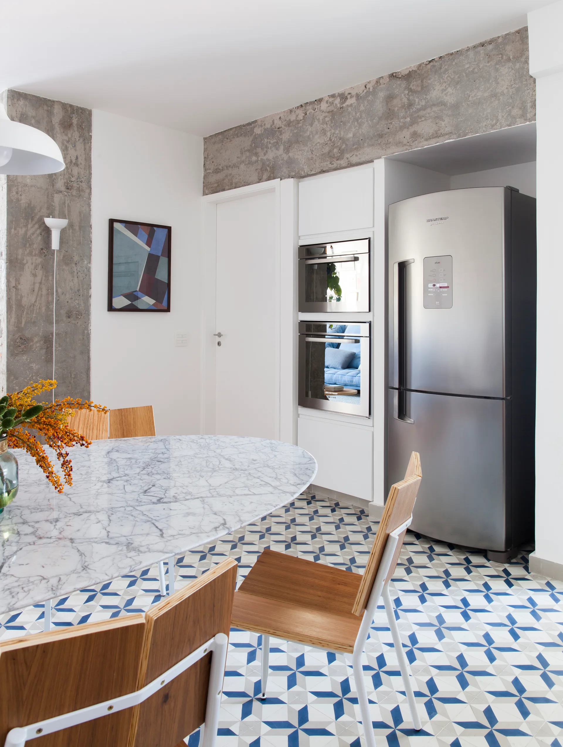 An oval marble dining table and wooden chairs stand on blue and white geometric tile flooring next to built-in kitchen appliances.