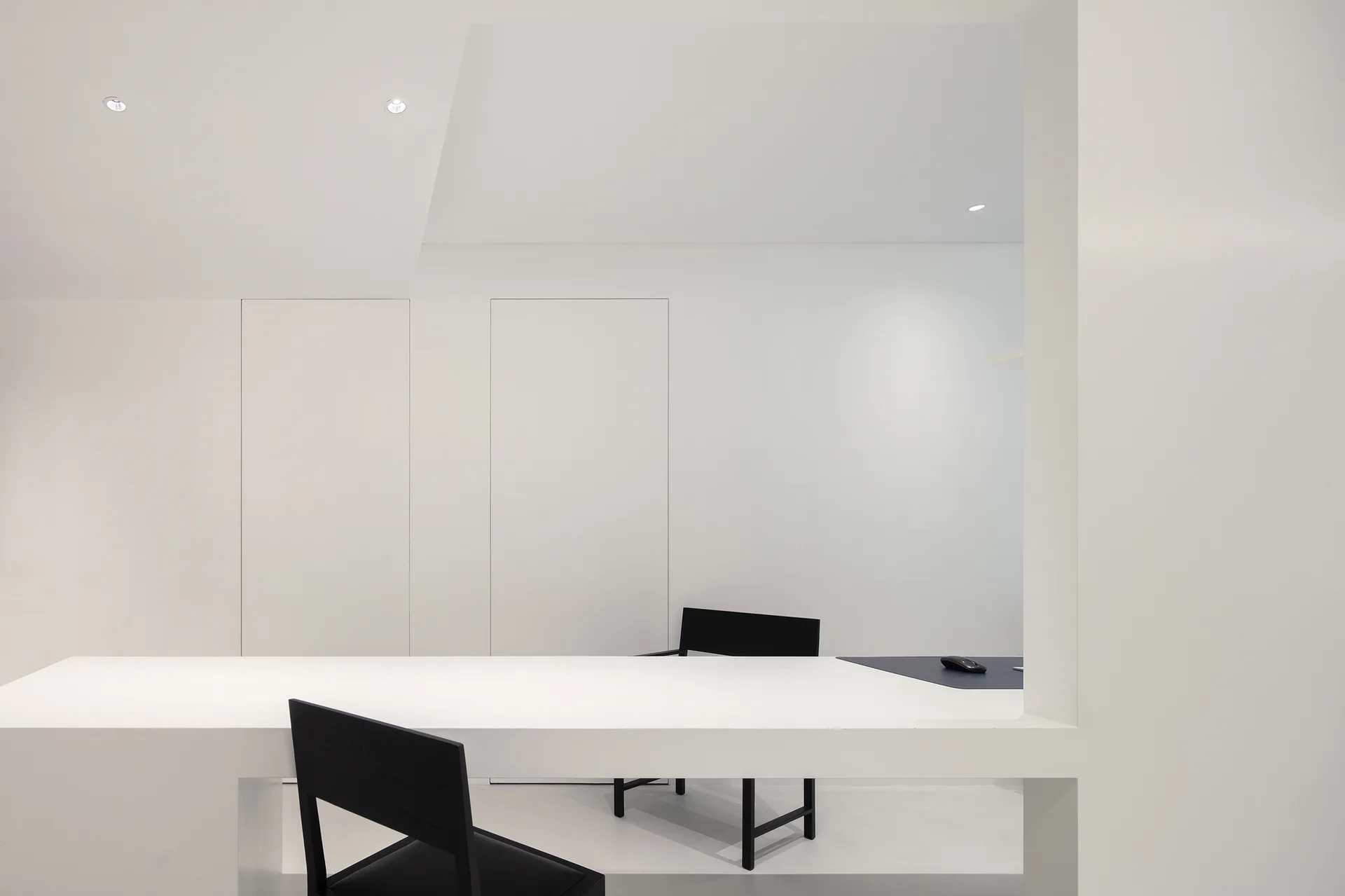A minimalist white office features a long white built-in desk, two simple black chairs, and seamless white wall panels.
