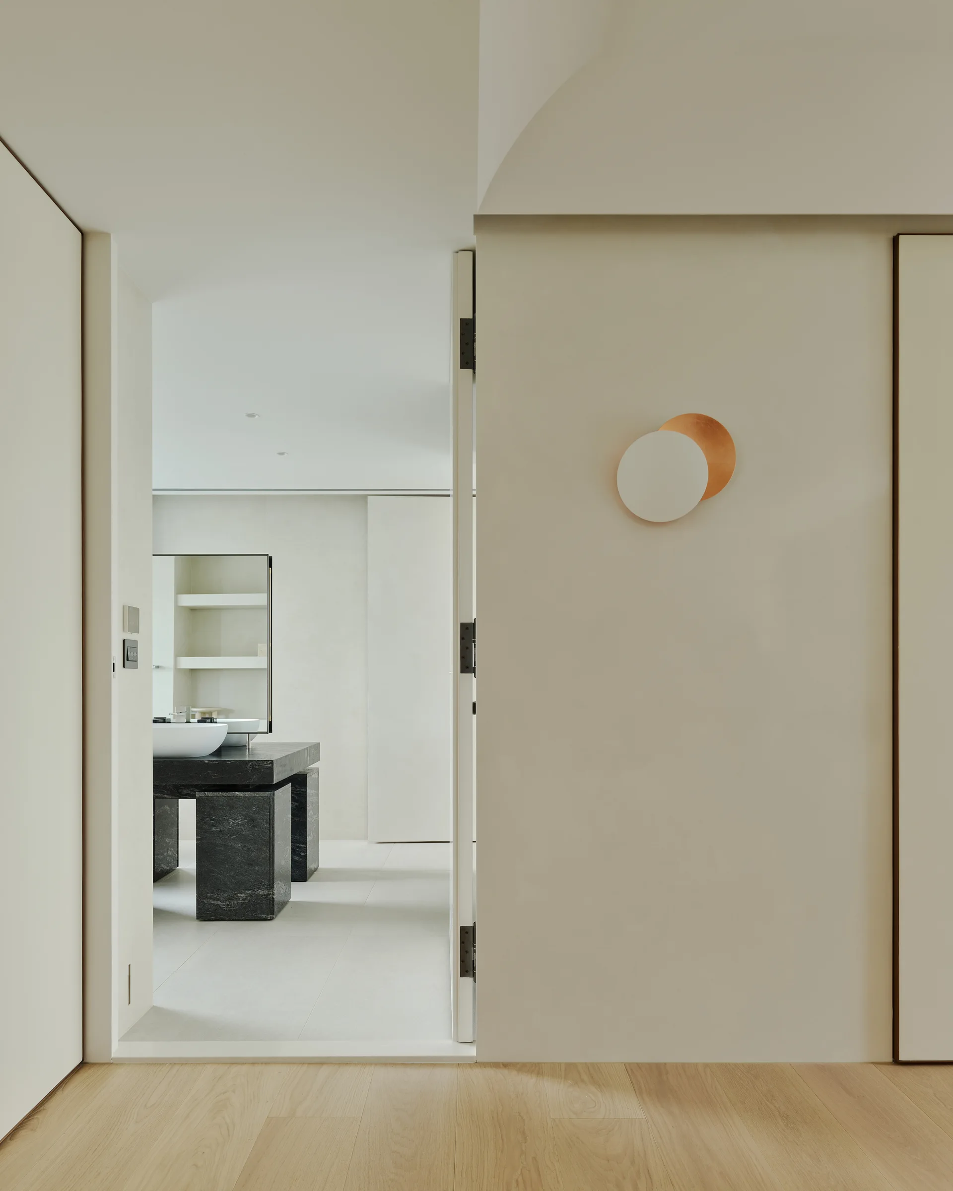 A view through a doorway into a minimalist bathroom featuring a dark stone vanity, with a circular copper wall sconce mounted on the foreground wall.