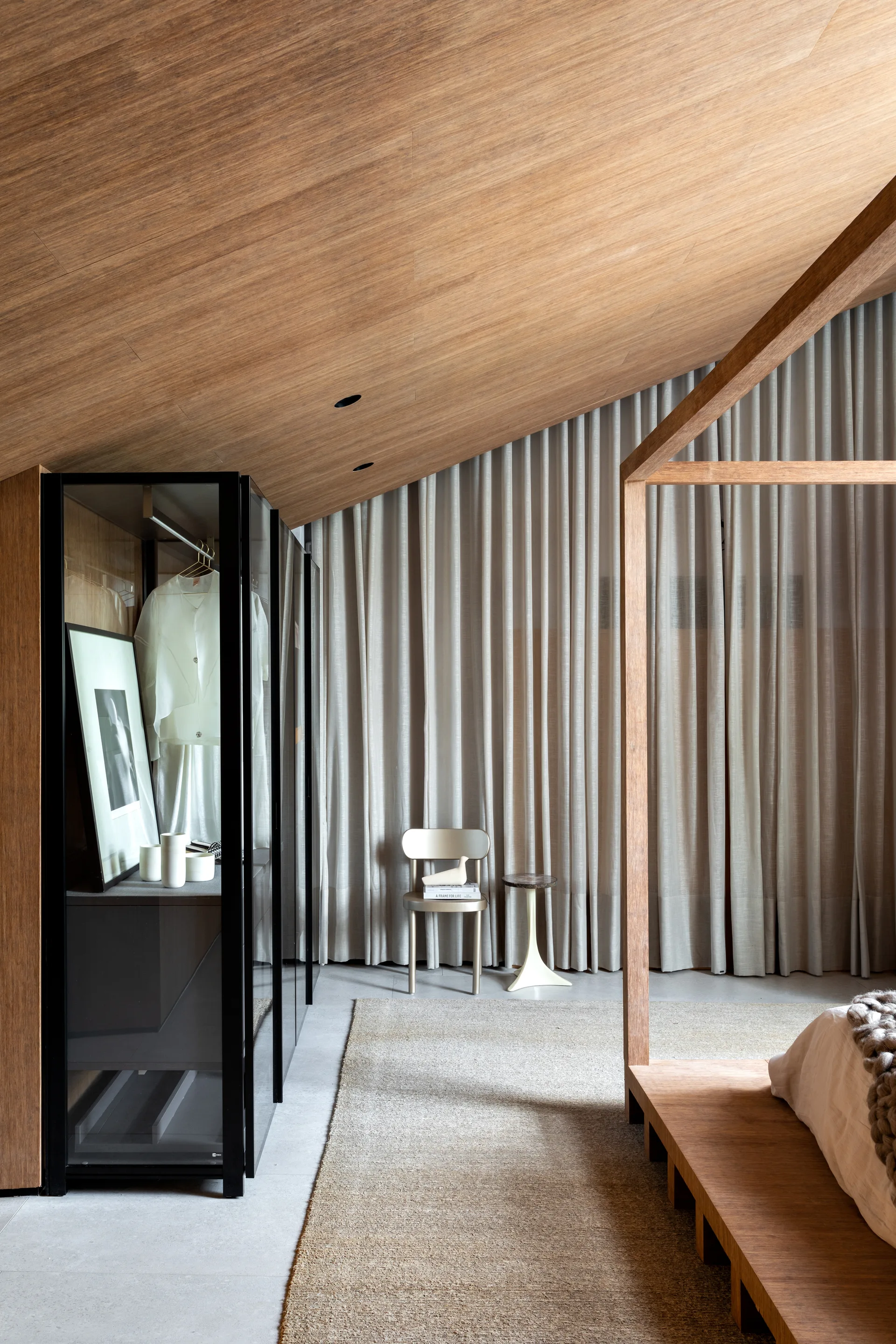 A modern bedroom featuring a sloped wood paneled ceiling, a wooden canopy bed frame, a black framed glass wardrobe, and floor to ceiling beige curtains.
