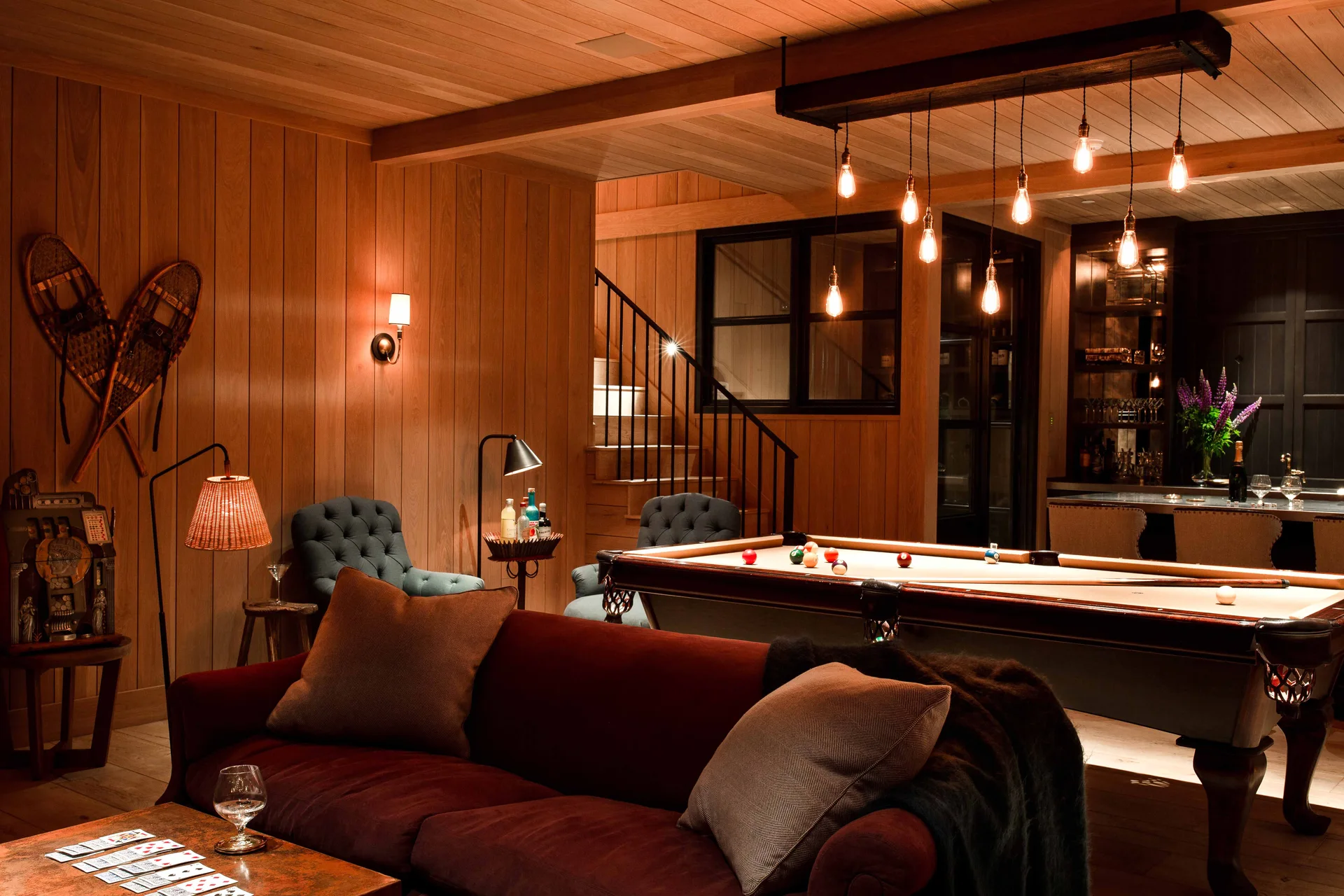 A rustic basement game room with wood paneling on the walls and ceiling, a pool table, and a burgundy velvet sofa.