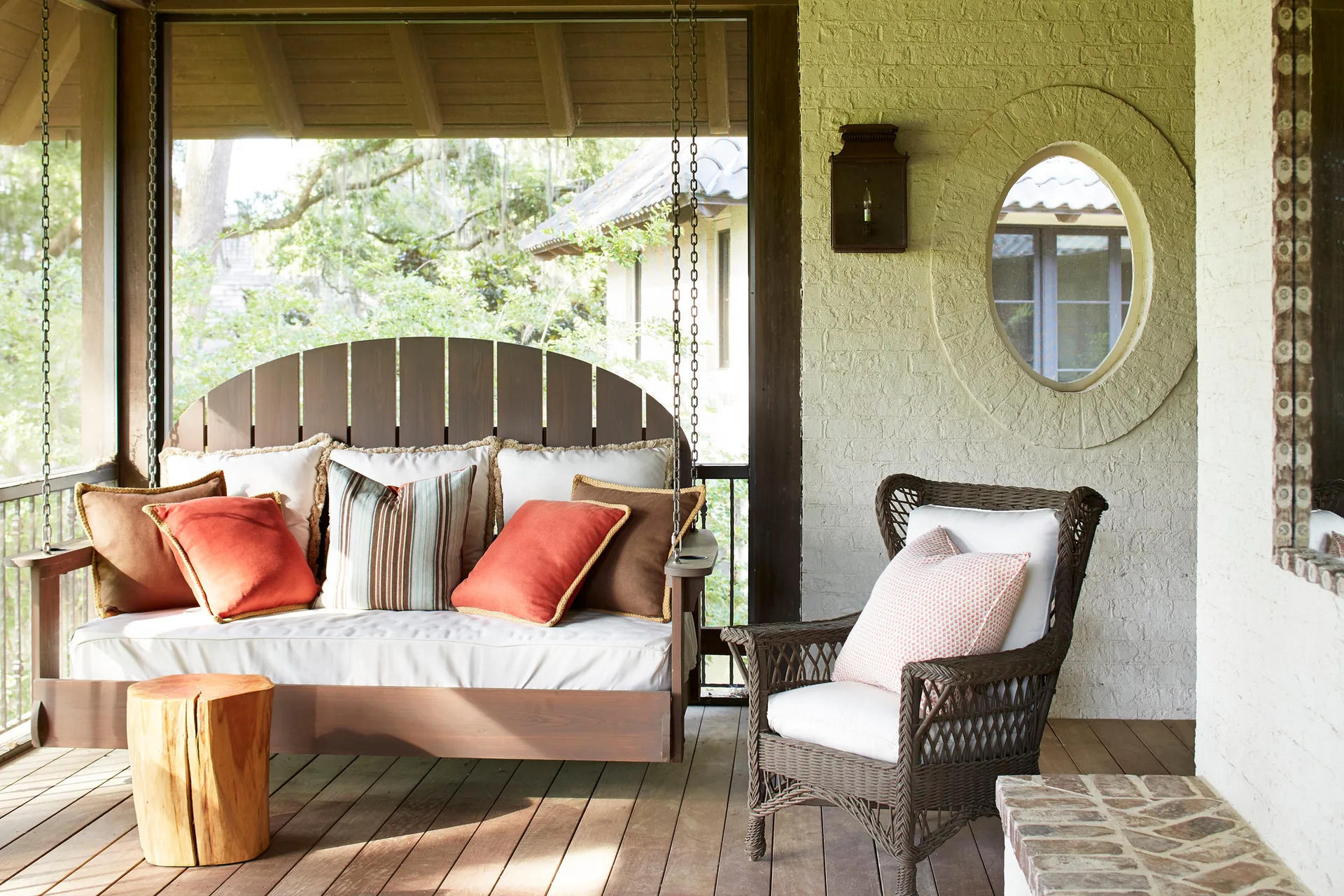 A covered porch featuring a wooden hanging swing and a wicker armchair on a plank deck next to a cream painted brick wall with a circular window.