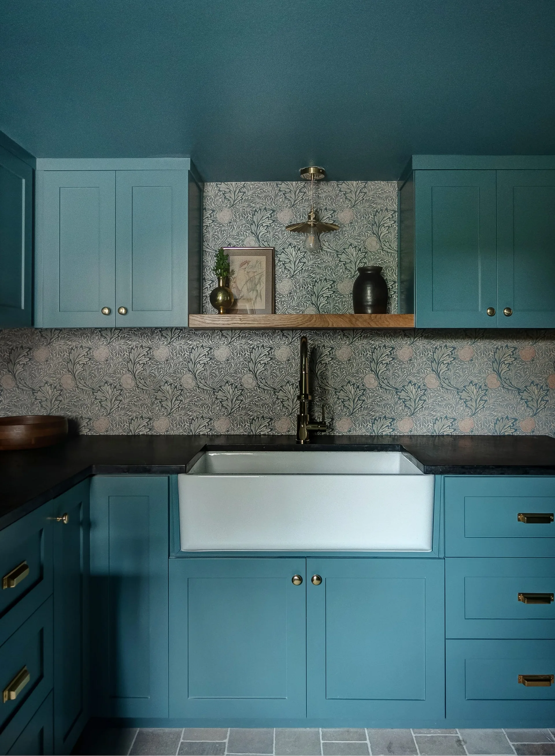 A kitchen featuring dark teal shaker cabinets, a botanical patterned wallpaper backsplash, a white farmhouse sink, and a floating wood shelf.
