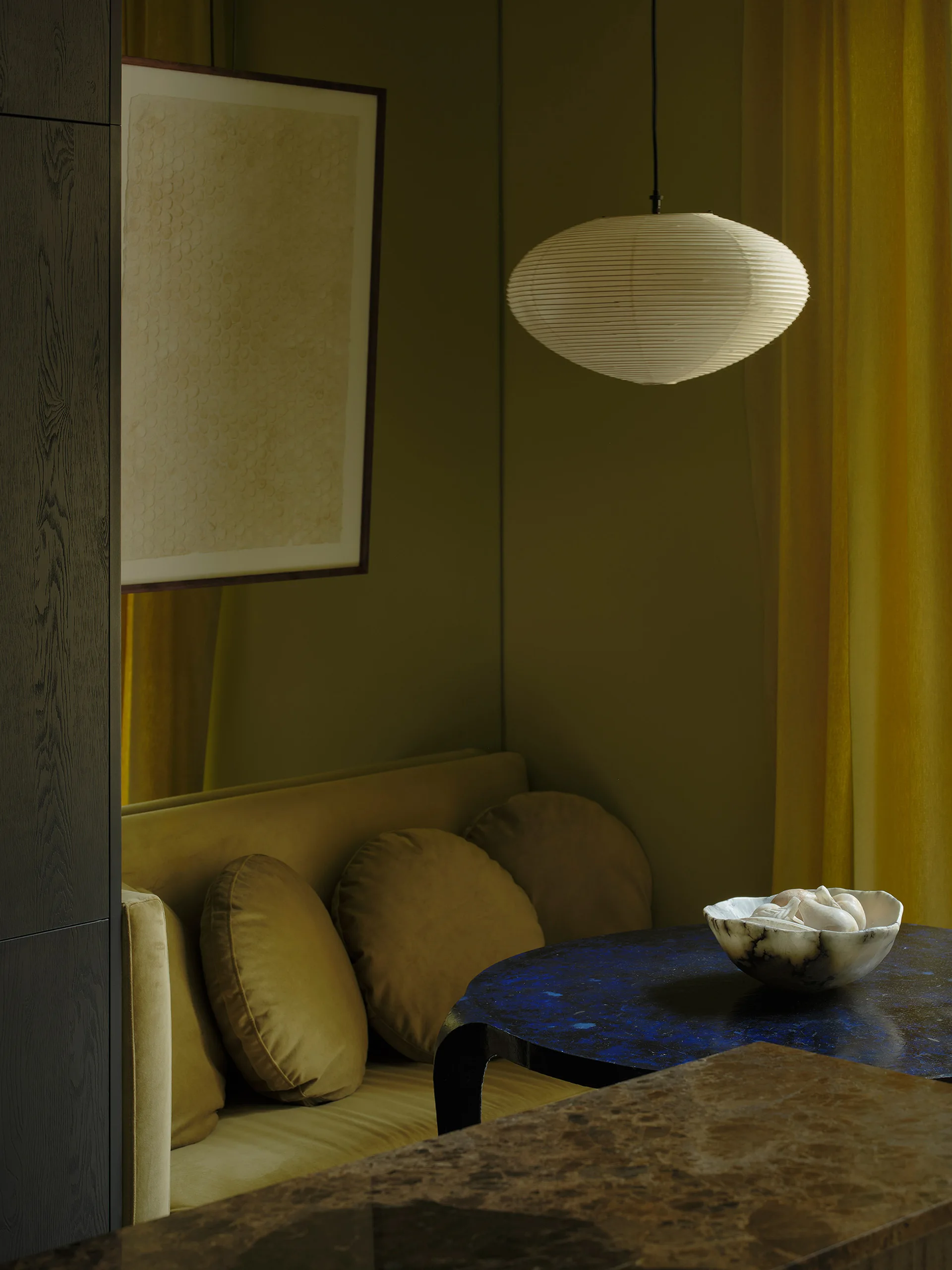 A dim corner seating area features a mustard yellow velvet sofa with round pillows, a circular blue table, and a glowing white paper pendant light.