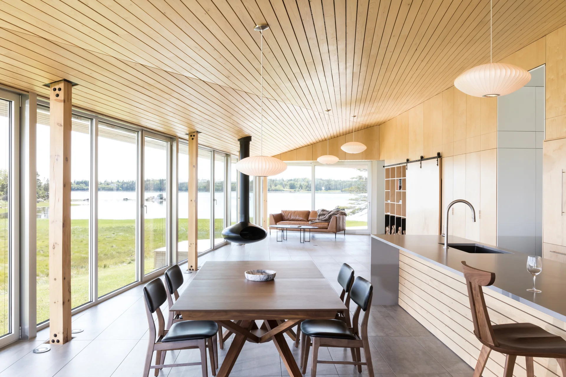 An open-concept dining and living area features a sloped wood ceiling, large windows overlooking water, a suspended fireplace, and a dining table.