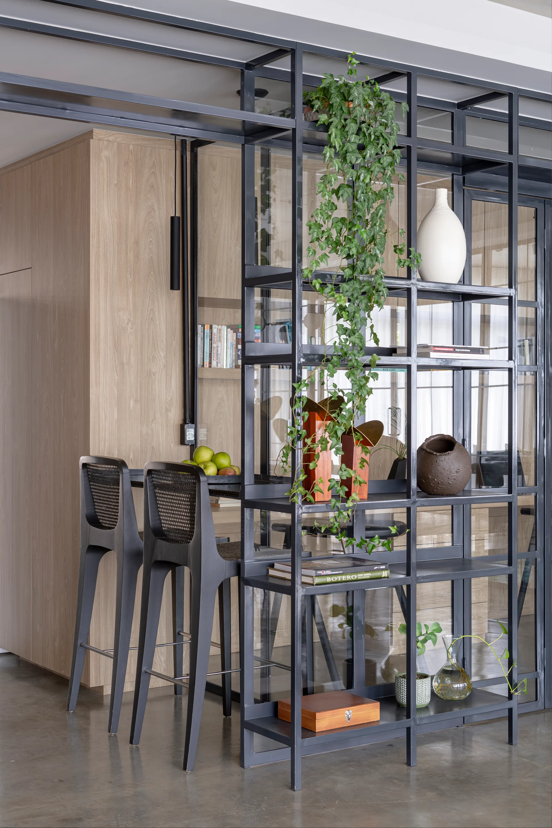A black metal grid shelving unit acts as a room divider next to two black cane-back bar stools against a light wood-paneled wall.