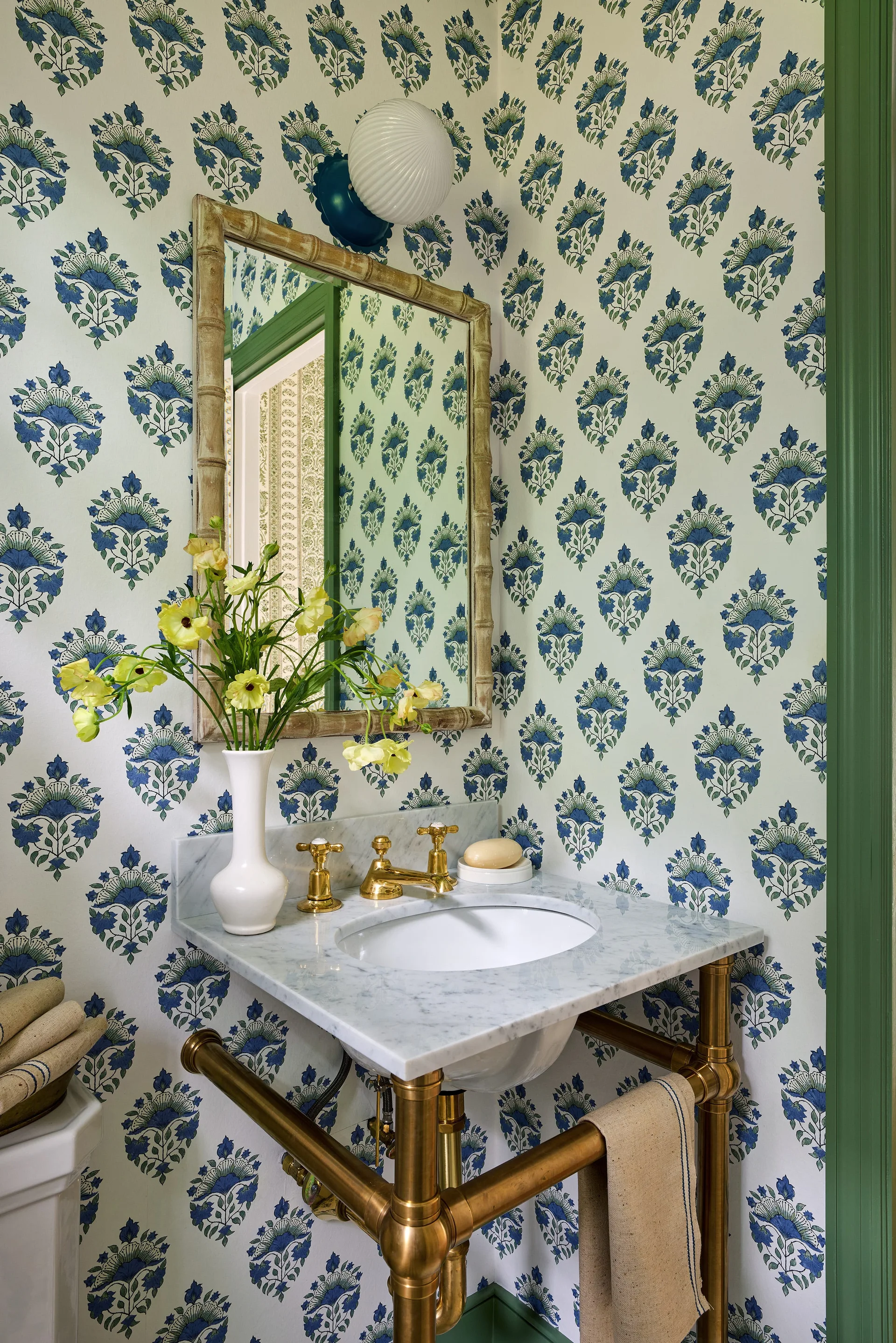 A small powder room features a marble vanity with exposed brass legs, floral wallpaper, and a bamboo-style mirror.