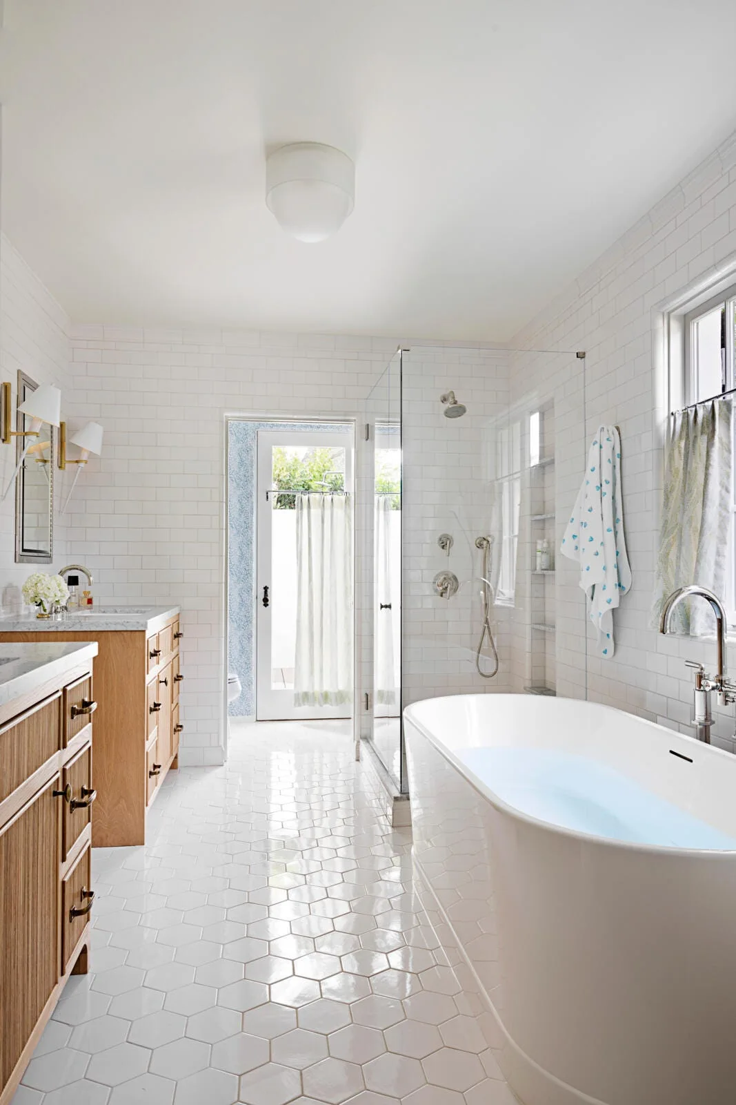 A bright transitional bathroom featuring white subway wall tiles, hexagonal floor tiles, a freestanding white tub, and a natural wood vanity.