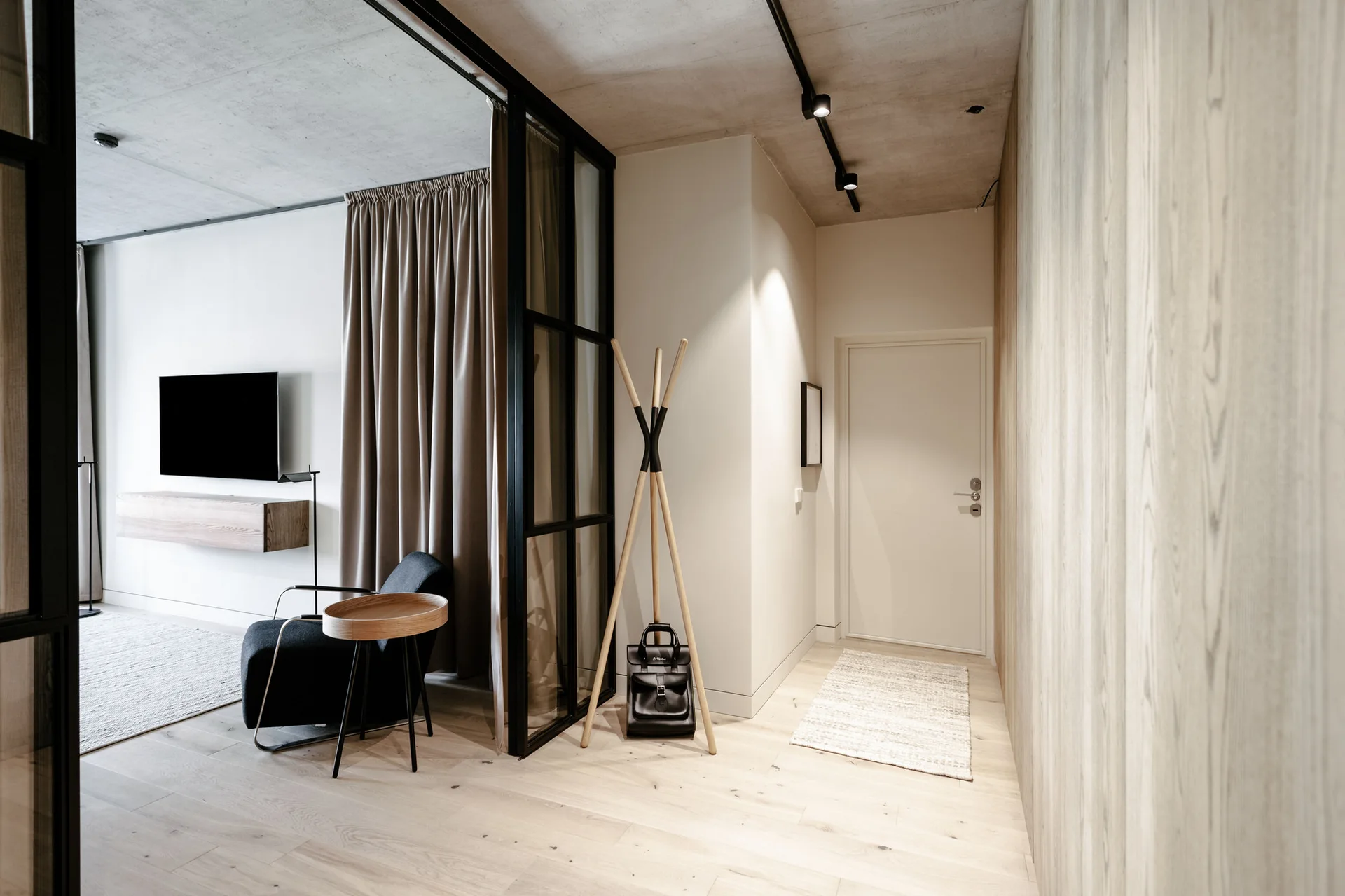 A modern entryway with light wood flooring, a vertical wood-paneled wall, and a black-framed glass partition separating it from a living room.