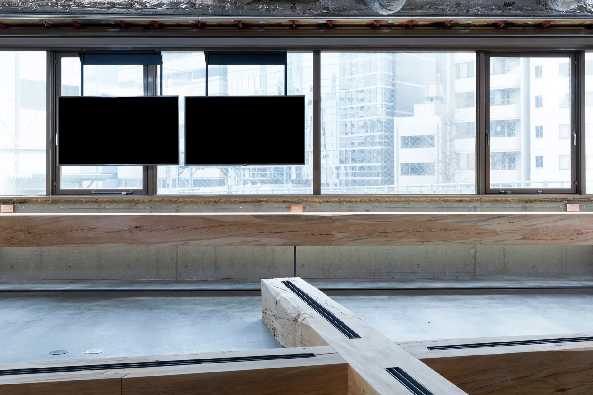 An industrial interior features two black screens suspended in front of large windows, raw concrete flooring, and thick light wood benches.