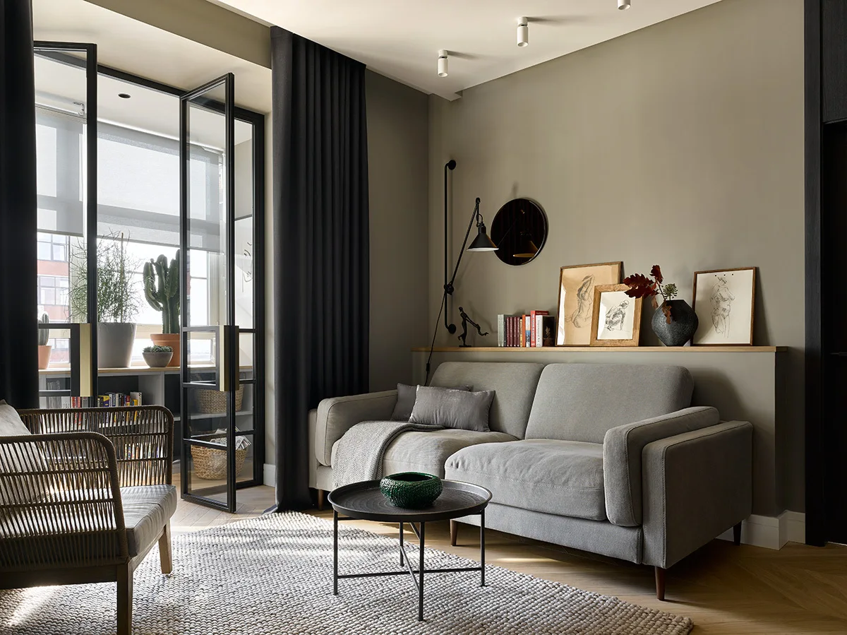A contemporary living room features a grey sofa against a display ledge, a round coffee table, and black-framed glass doors leading to a balcony.
