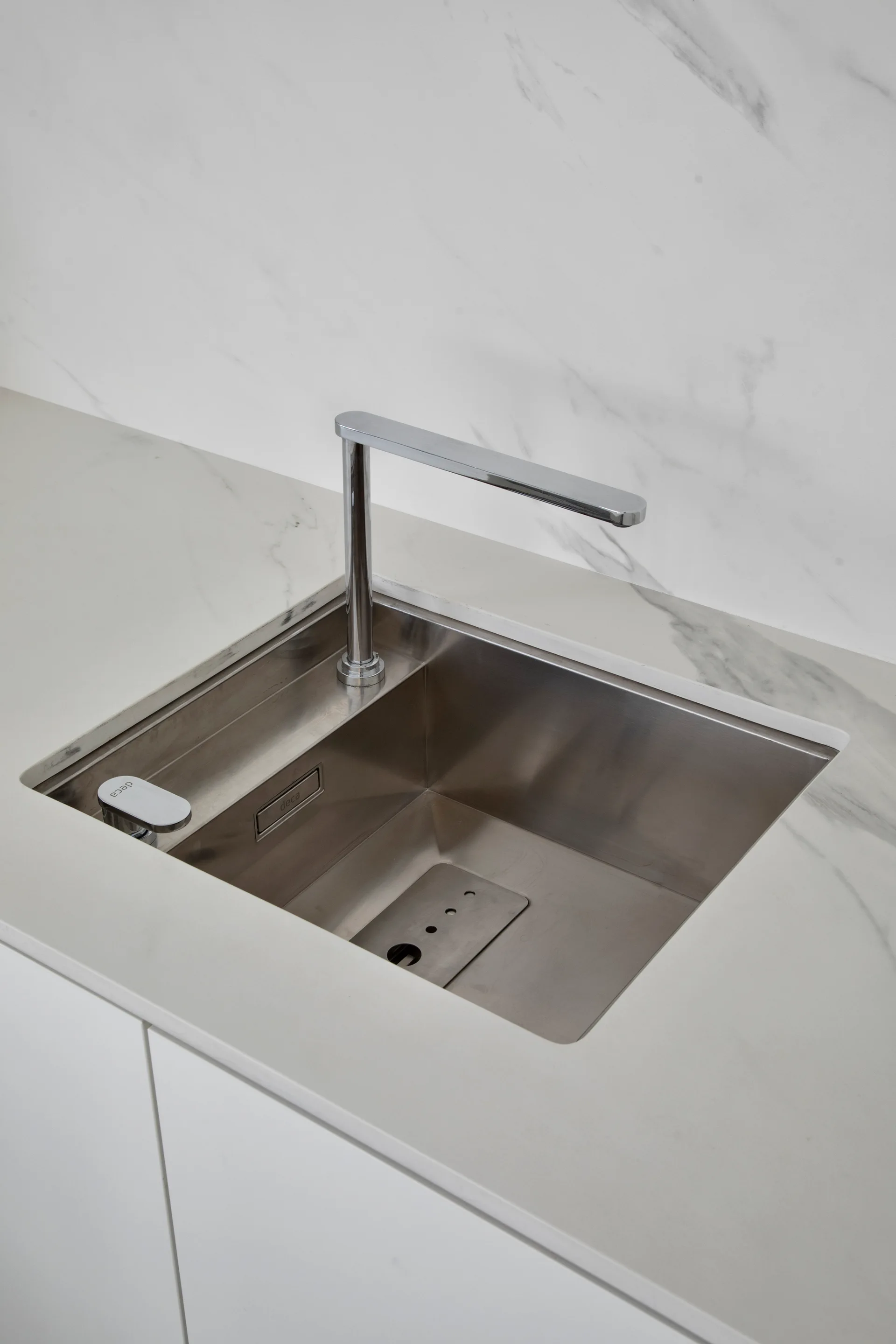 A minimalist stainless steel undermount kitchen sink with a sleek chrome faucet, set into a veined white marble countertop and matching backsplash.