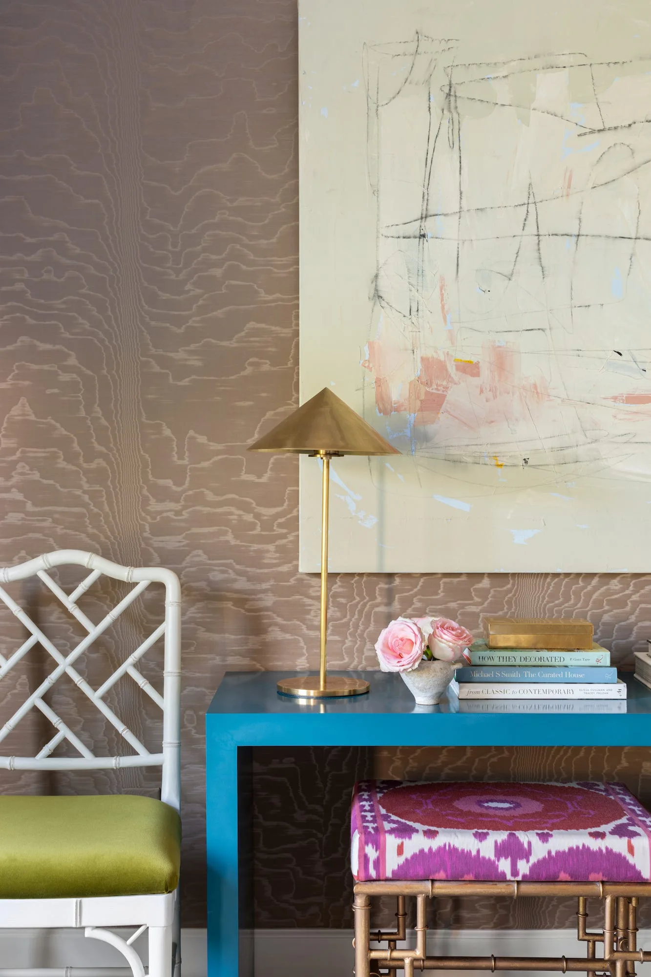 A bright blue console table with a brass table lamp and stacked books sits against organic wood-grain wallpaper and a white lattice chair.