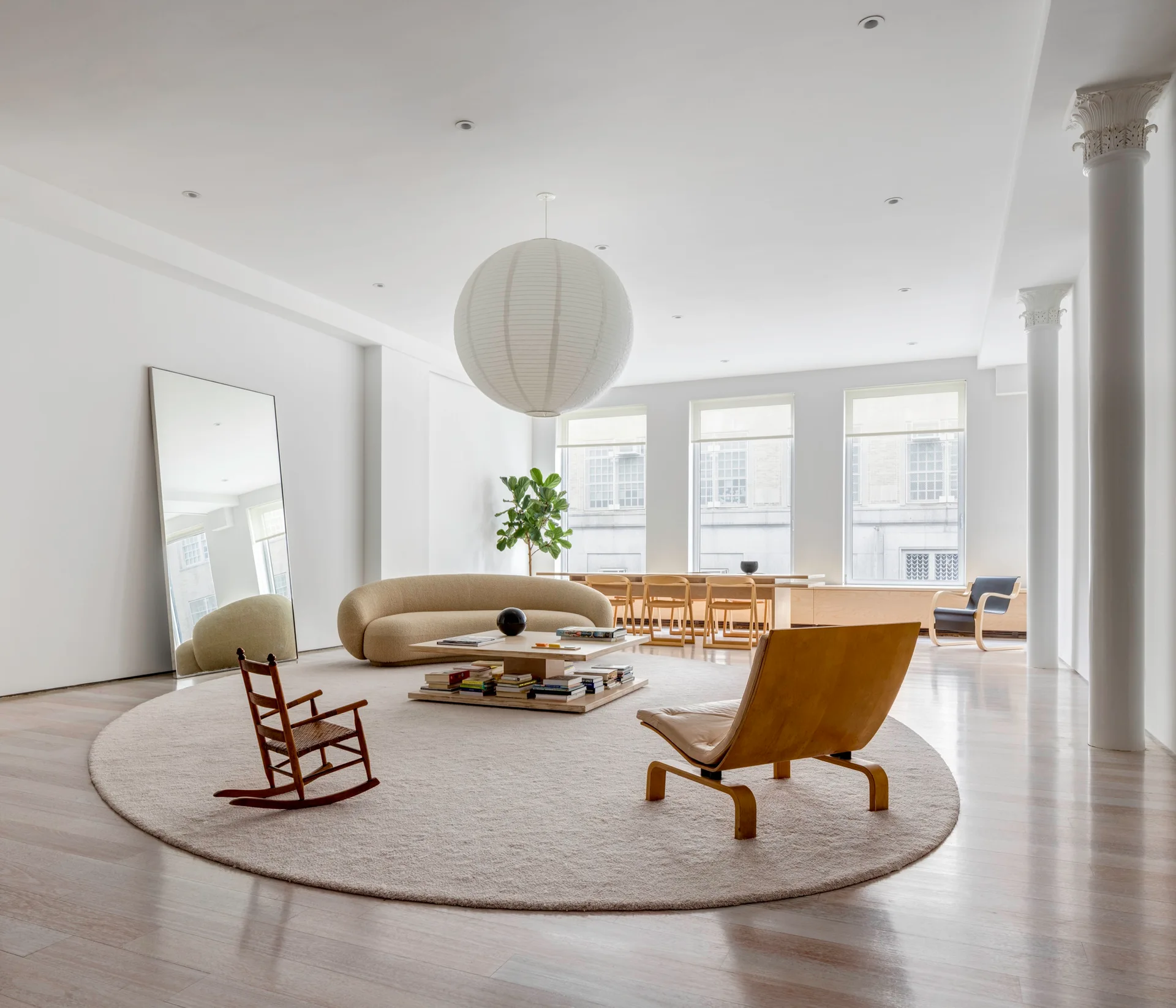 A spacious white living room features a large round rug, a curved beige sofa, a low wooden coffee table, and an oversized spherical paper pendant light.