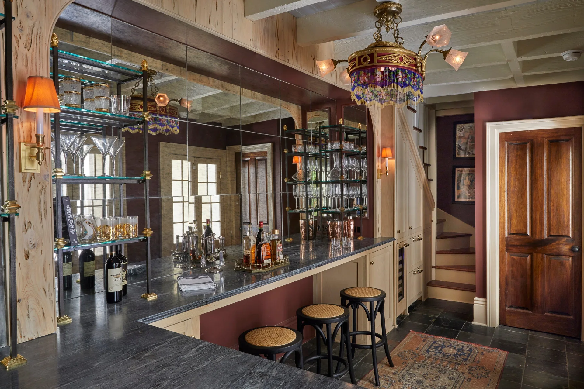 A dark burgundy home bar features a stone-topped counter, a grid-patterned mirrored wall, three cane-seated stools, and an ornate vintage chandelier.