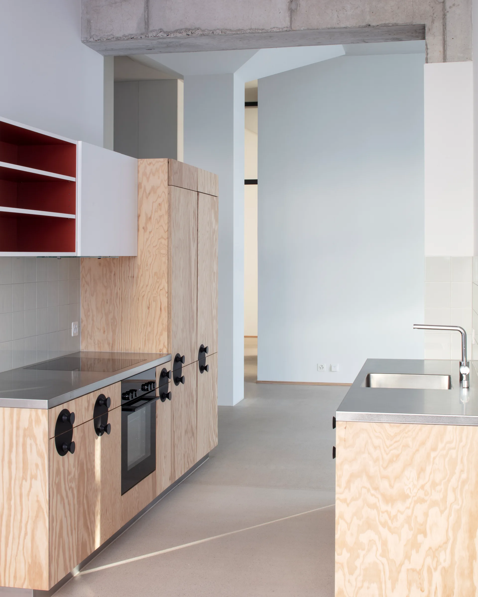 A minimalist kitchen features plywood cabinetry with oversized black handles, stainless steel countertops, a sink, and an exposed concrete beam overhead.