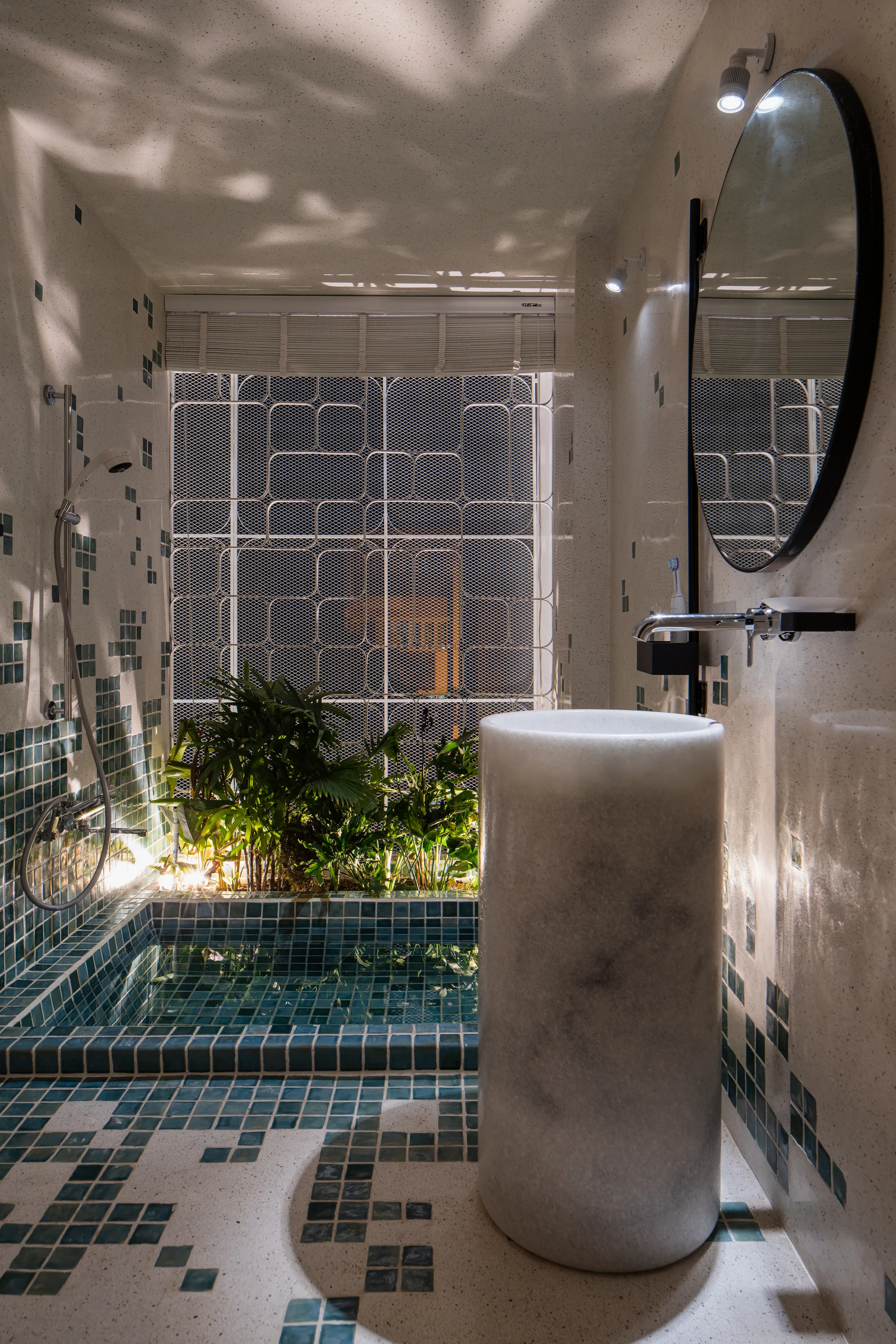 A contemporary bathroom featuring a tall cylindrical stone pedestal sink, a circular mirror, and a sunken shower lined with teal mosaic tiles.