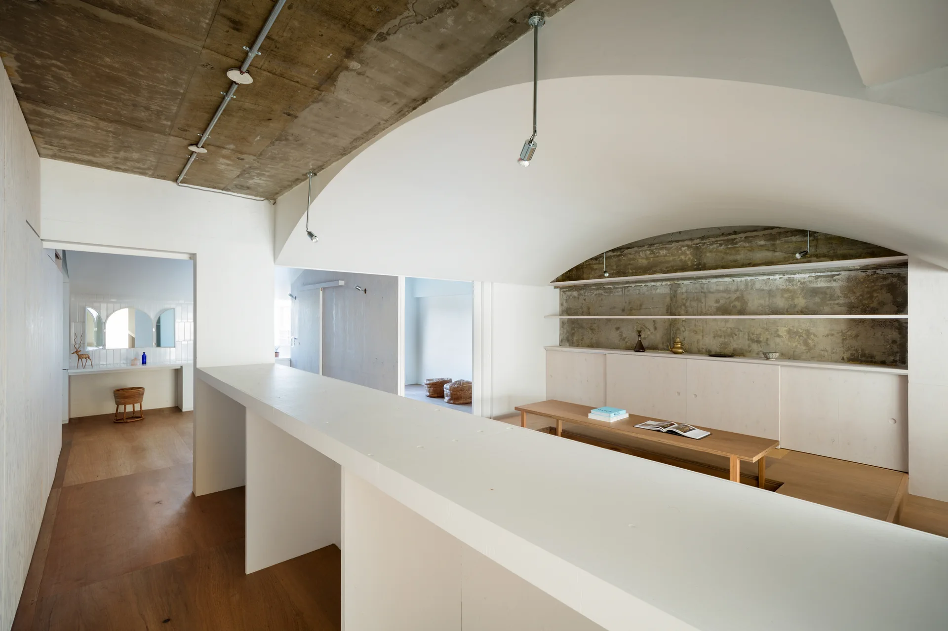 An open-plan living area features a dramatic curved white ceiling set against a raw concrete ceiling, complemented by light wood cabinets and a low bench.
