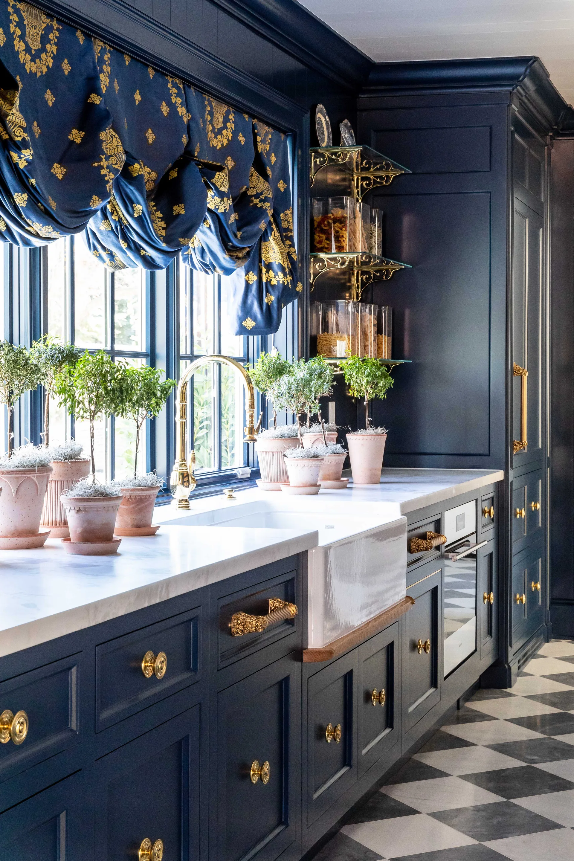 A traditional navy blue kitchen features a white apron sink, marble countertops, brass hardware, and a black and white checkered floor.