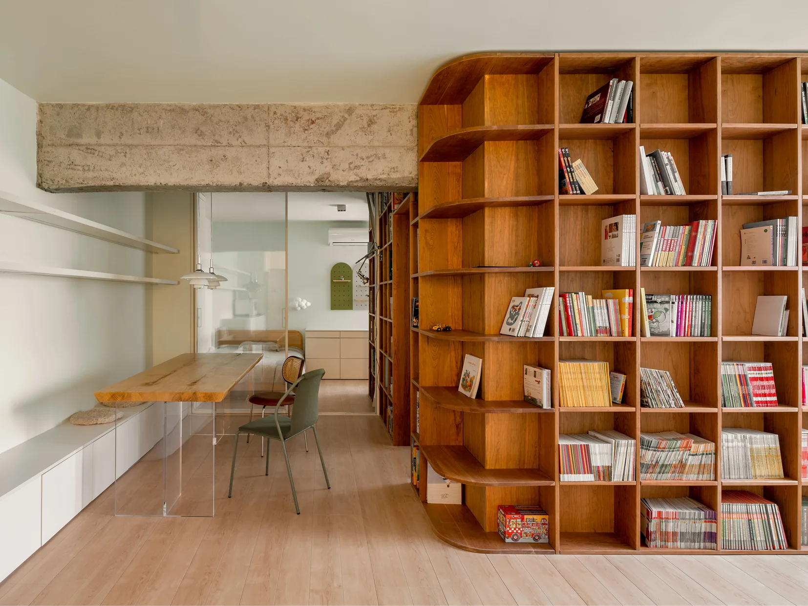 A modern interior featuring a large curved wooden bookshelf, a dining table with clear acrylic legs, and an exposed raw concrete beam.