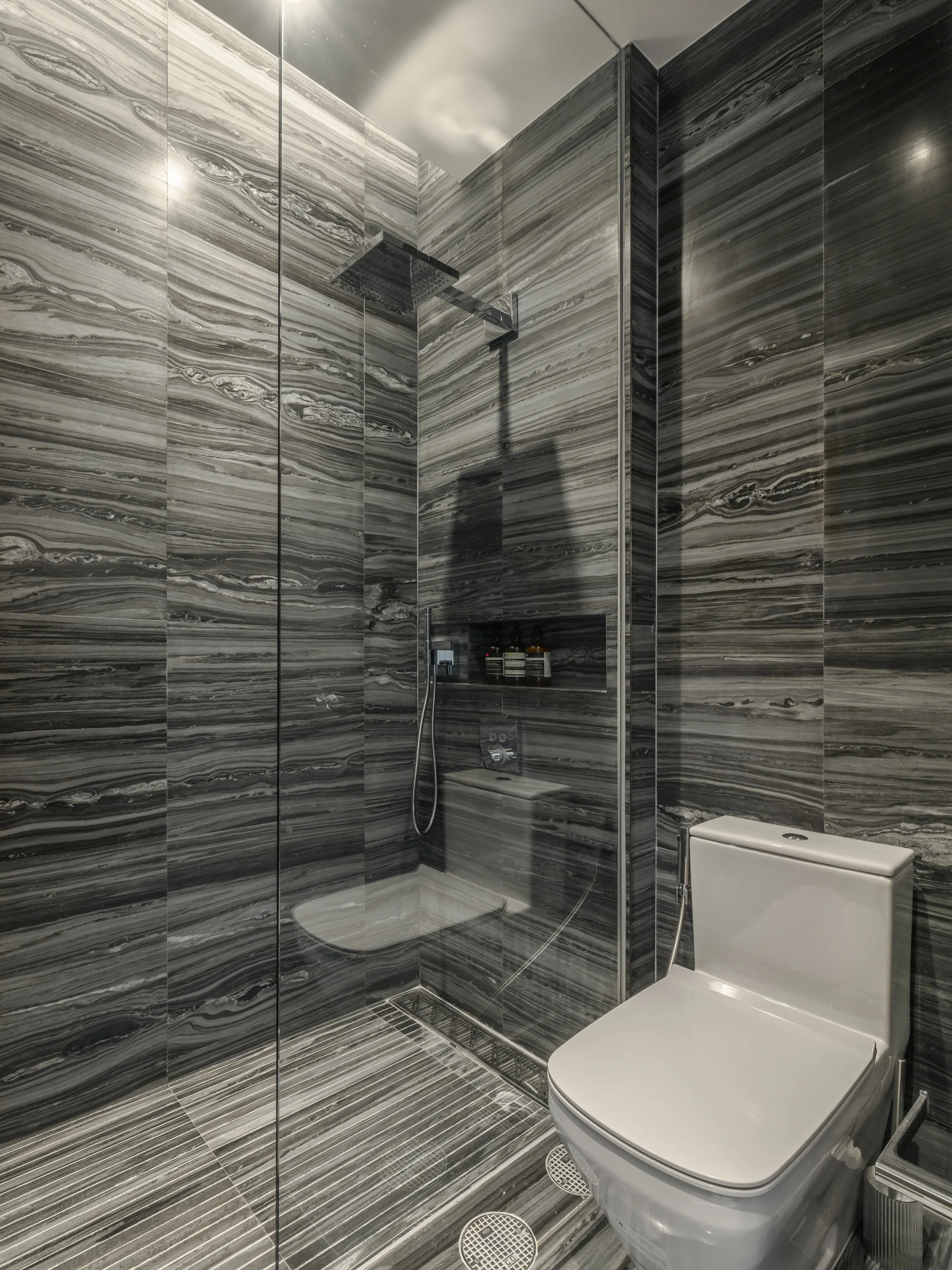 A modern bathroom featuring dark grey veined stone tile walls and flooring, a glass walk-in shower enclosure, and a white ceramic toilet.