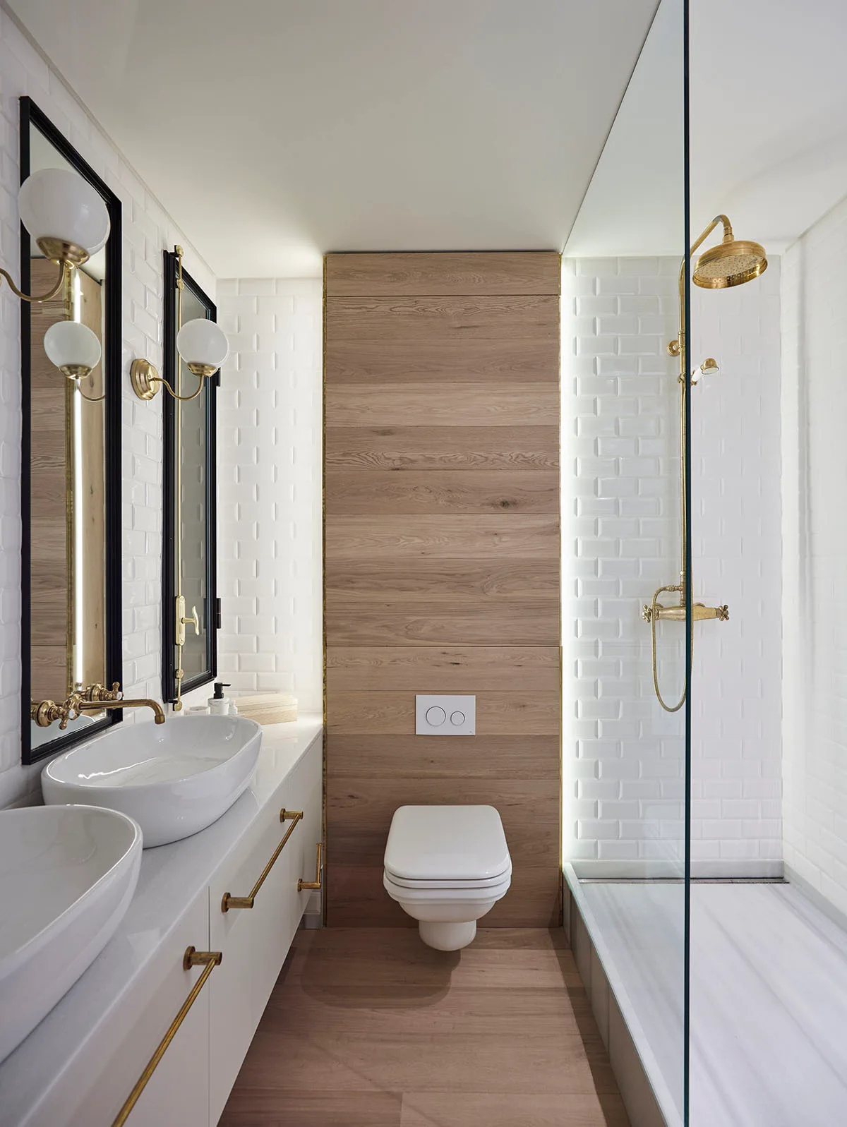 A modern bathroom with a double sink vanity, a wall-mounted toilet on a wood accent wall, and a walk-in shower with gold hardware.