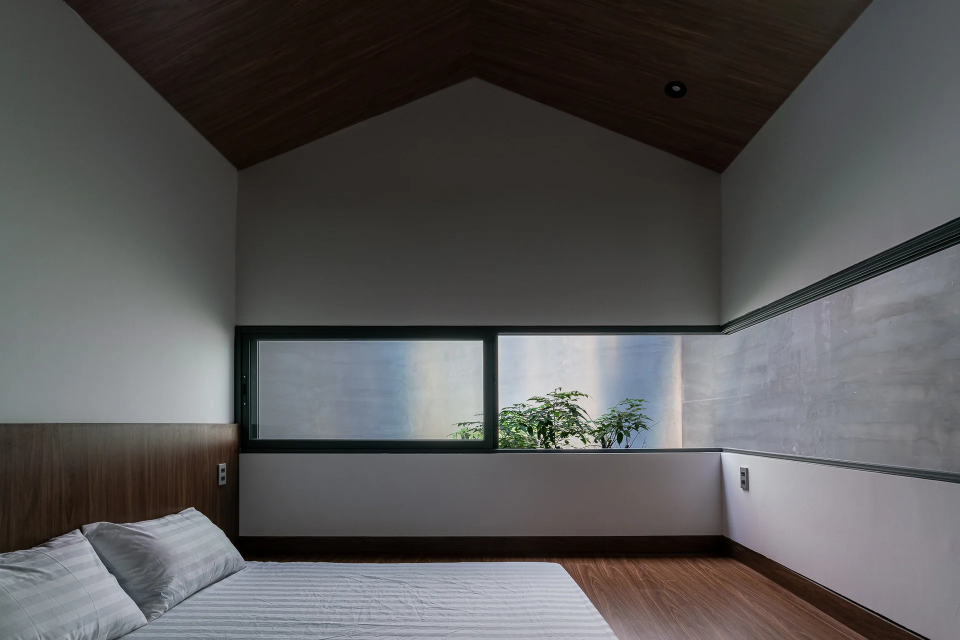 A minimalist bedroom featuring a bed with striped sheets, a vaulted wood ceiling, and a low horizontal corner window looking out at greenery.