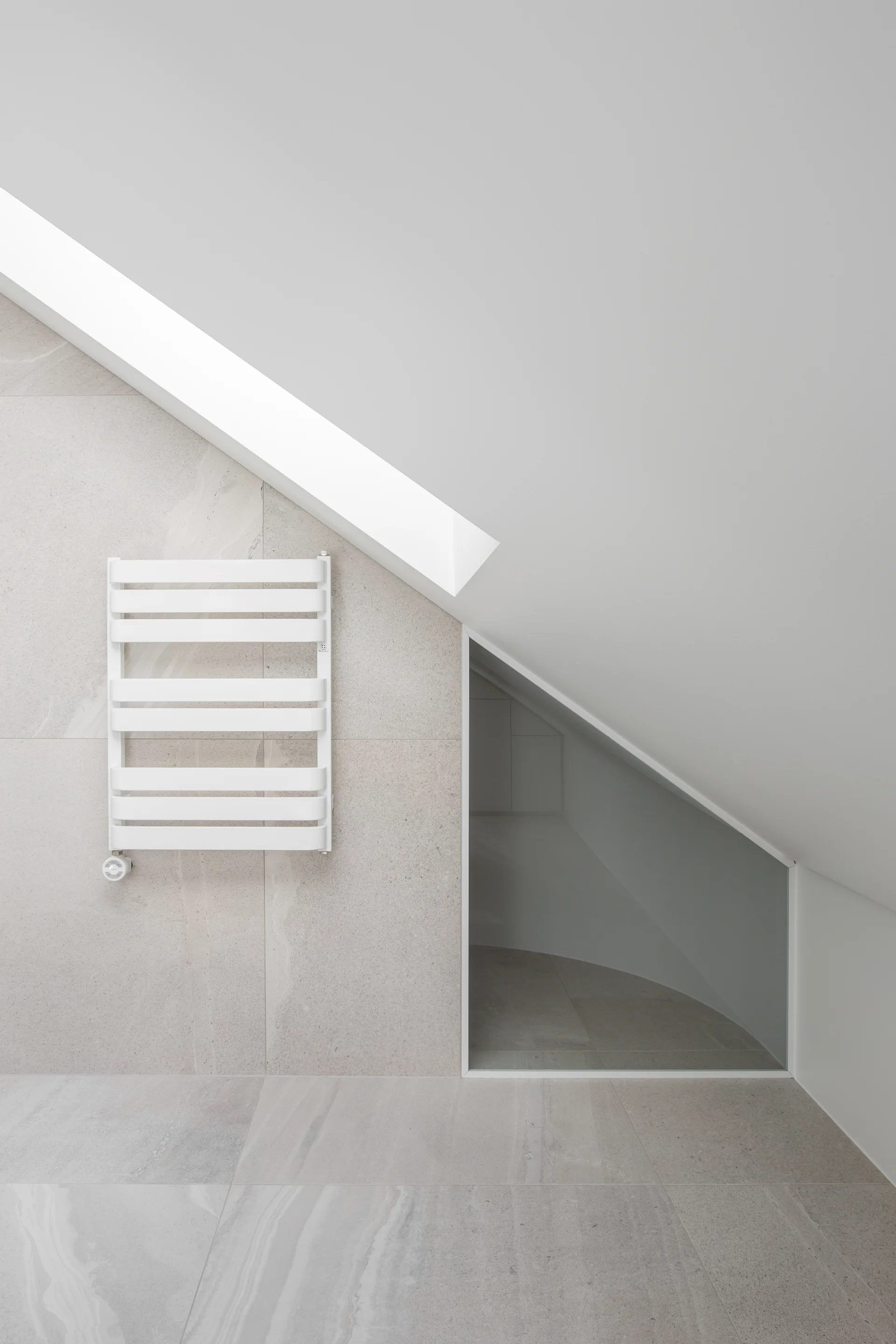 A white towel warmer is mounted on a light grey tiled wall under a sloped white ceiling in a minimalist attic bathroom.