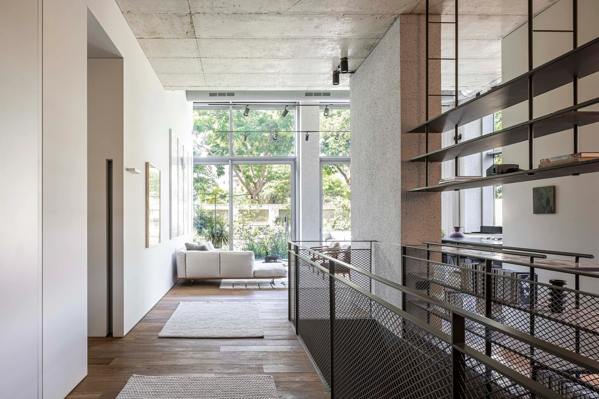 A modern mezzanine hallway with wooden floors, an exposed concrete ceiling, dark steel mesh railings, and open shelving.