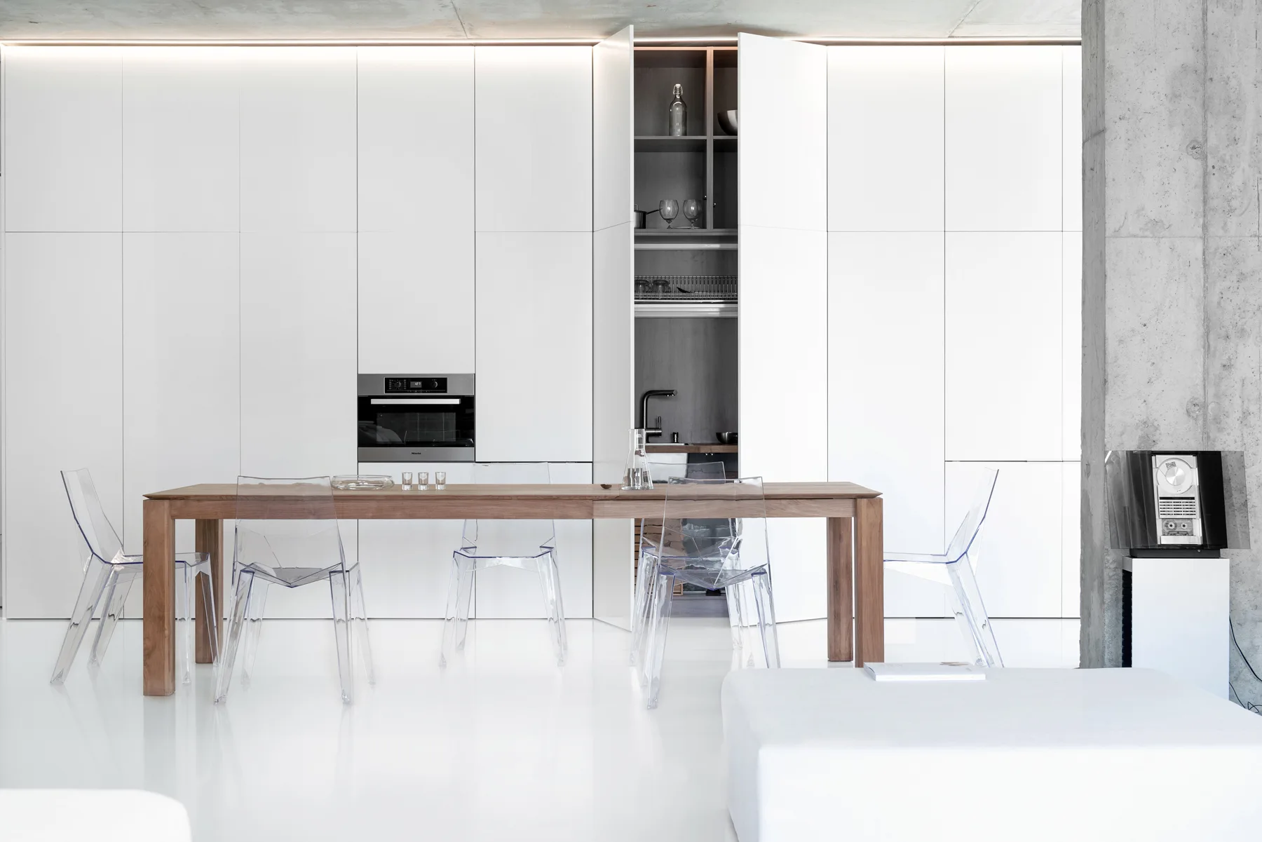 A minimalist white kitchen features a long wooden dining table and clear acrylic chairs in front of a seamless wall of cabinetry.