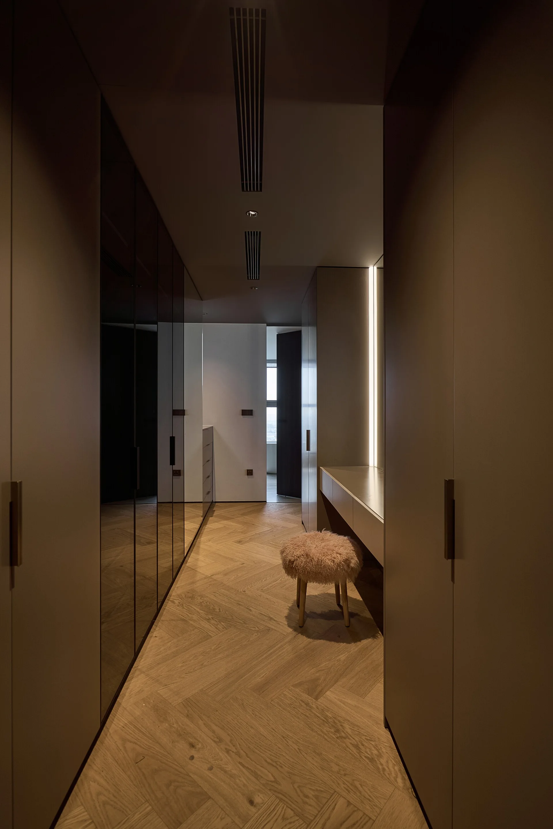 A modern dressing room featuring matte wardrobes, dark reflective panels, a built-in vanity with a textured stool, and herringbone wood flooring.