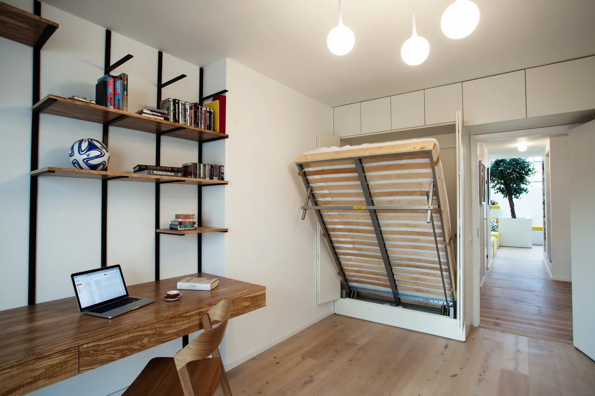 A modern white bedroom features a wooden desk with black metal shelving and a partially lowered murphy bed set into built-in cabinetry.