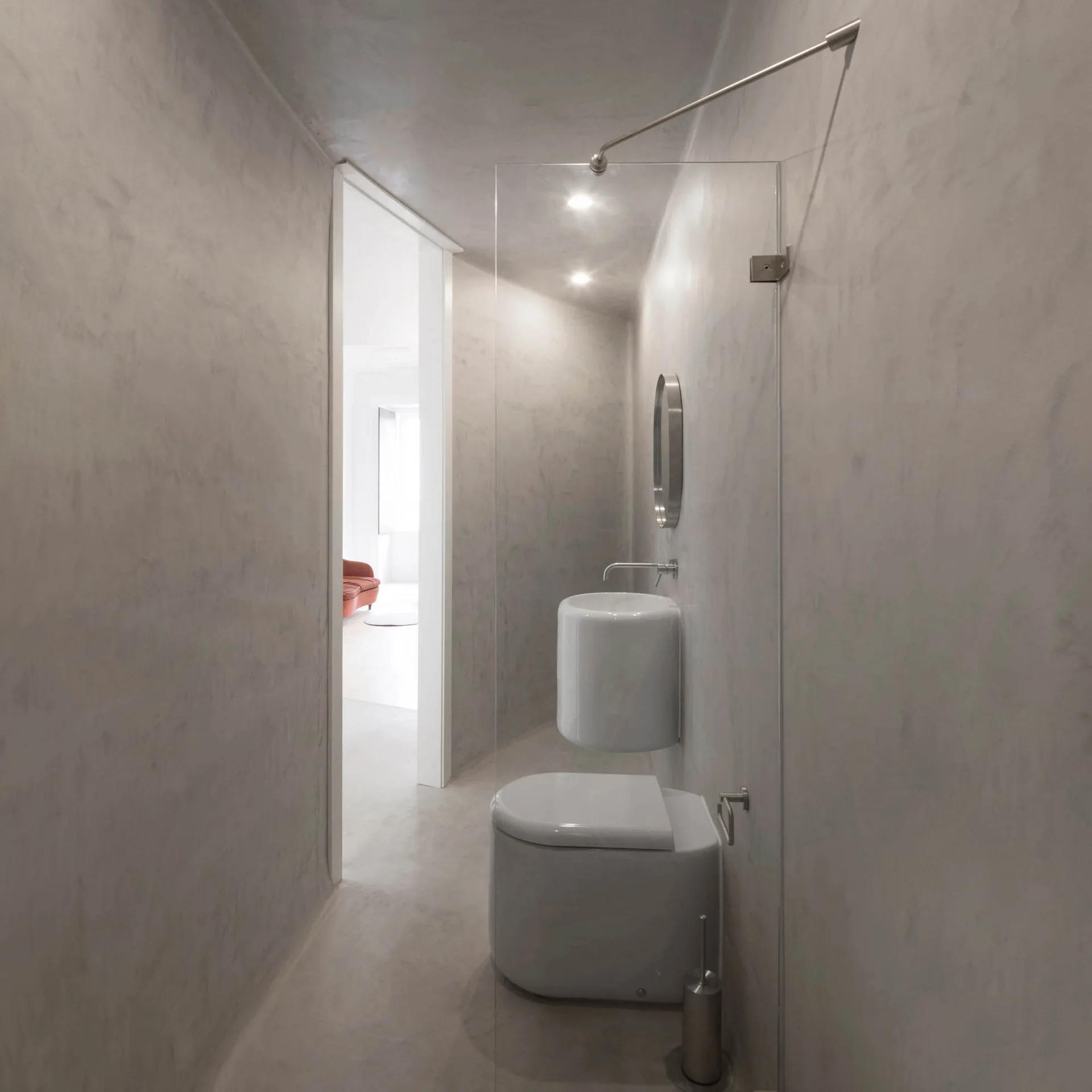 A minimalist bathroom features seamless gray plaster walls, a white wall-mounted sink, a toilet, and a glass shower partition.