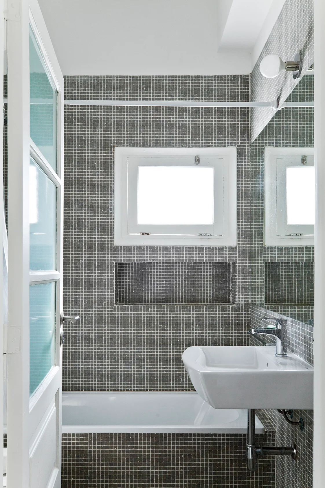 A compact bathroom featuring grey mosaic tiled walls, a white wall-mounted sink, a bathtub, and a recessed wall niche.