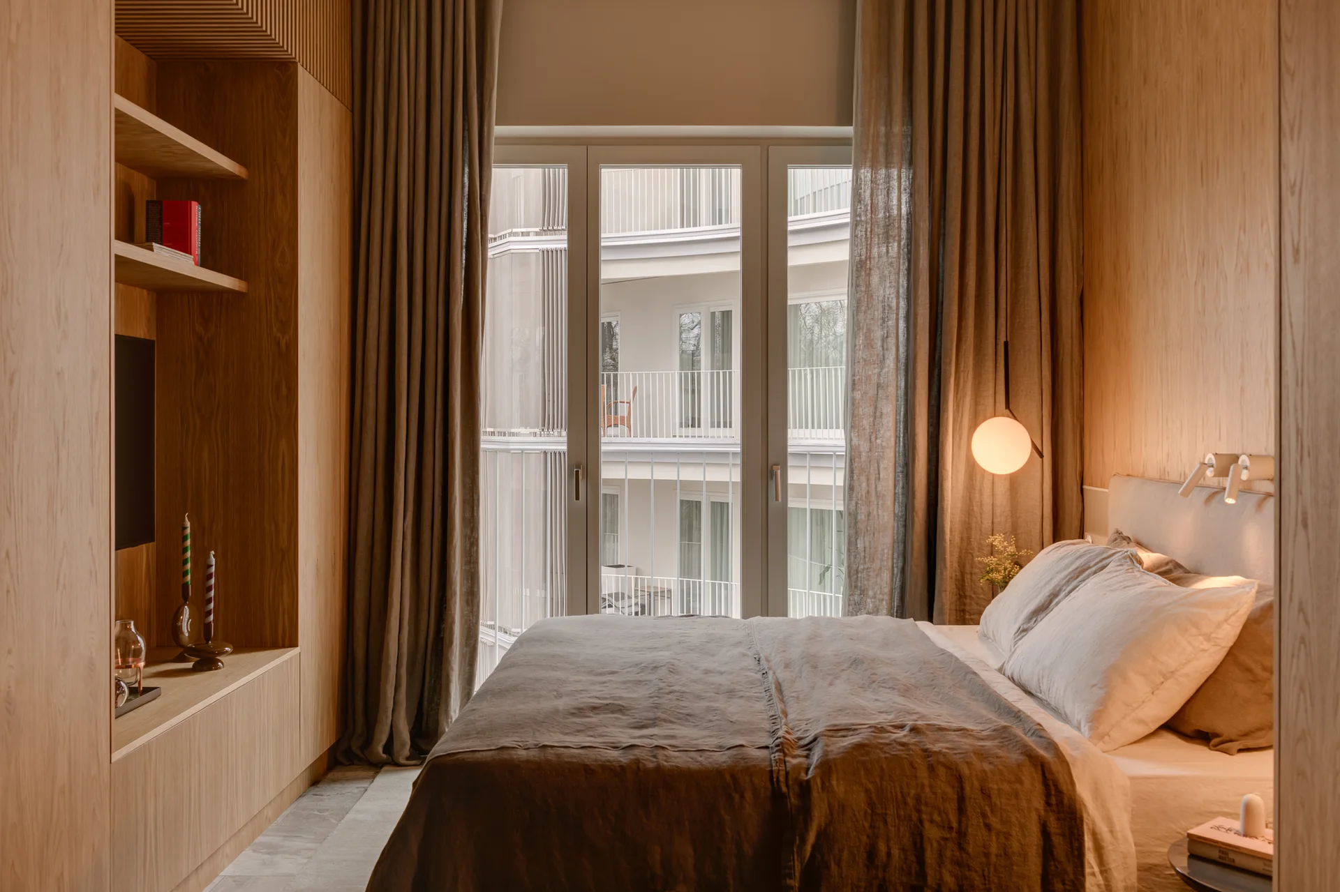 A modern bedroom features light wood wall paneling, a bed with linen sheets, a globe pendant light, and floor-length curtains framing a window.
