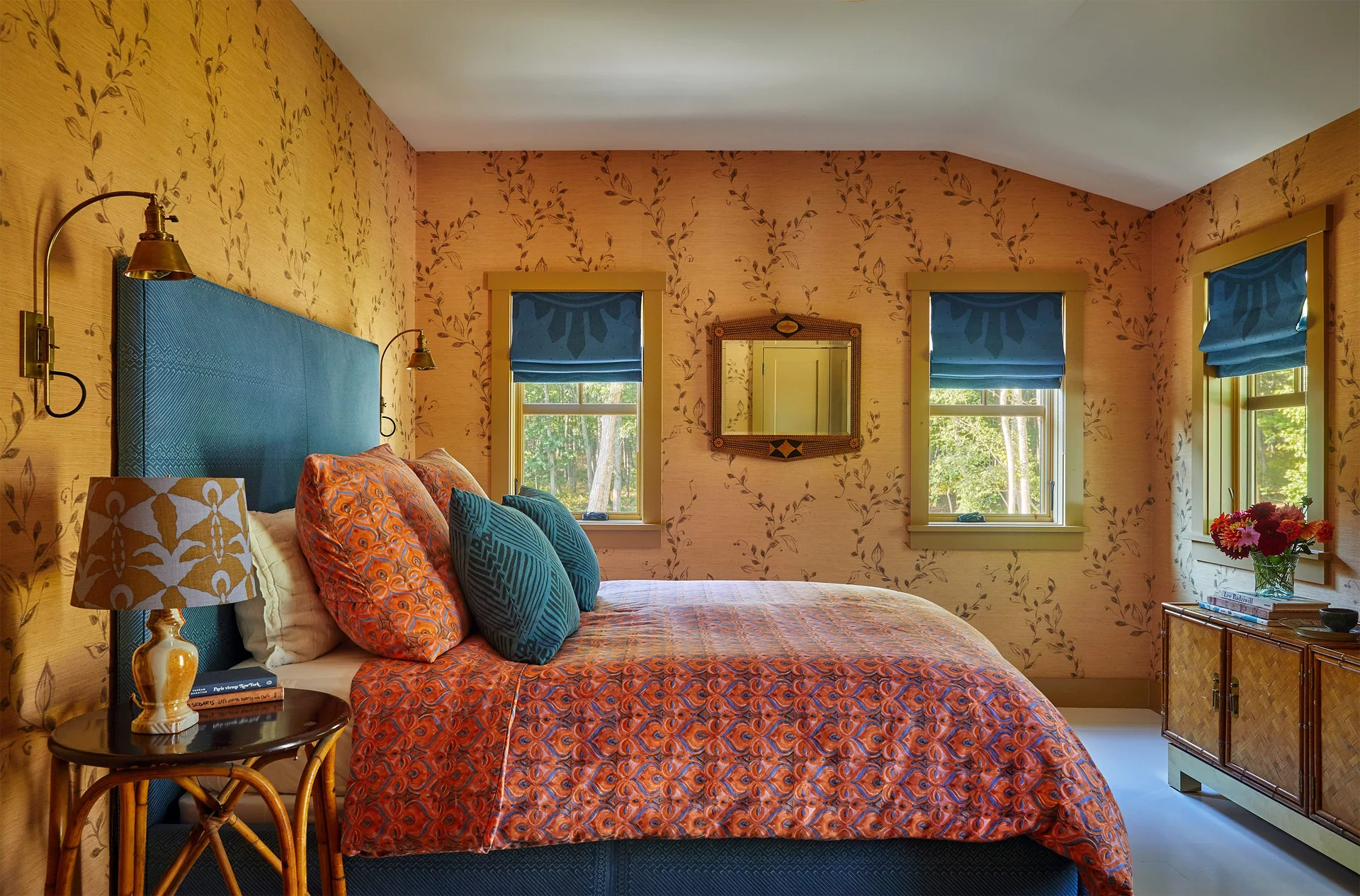 An eclectic bedroom features warm botanical wallpaper, a tall blue upholstered headboard, and matching blue roman shades over wood-trimmed windows.