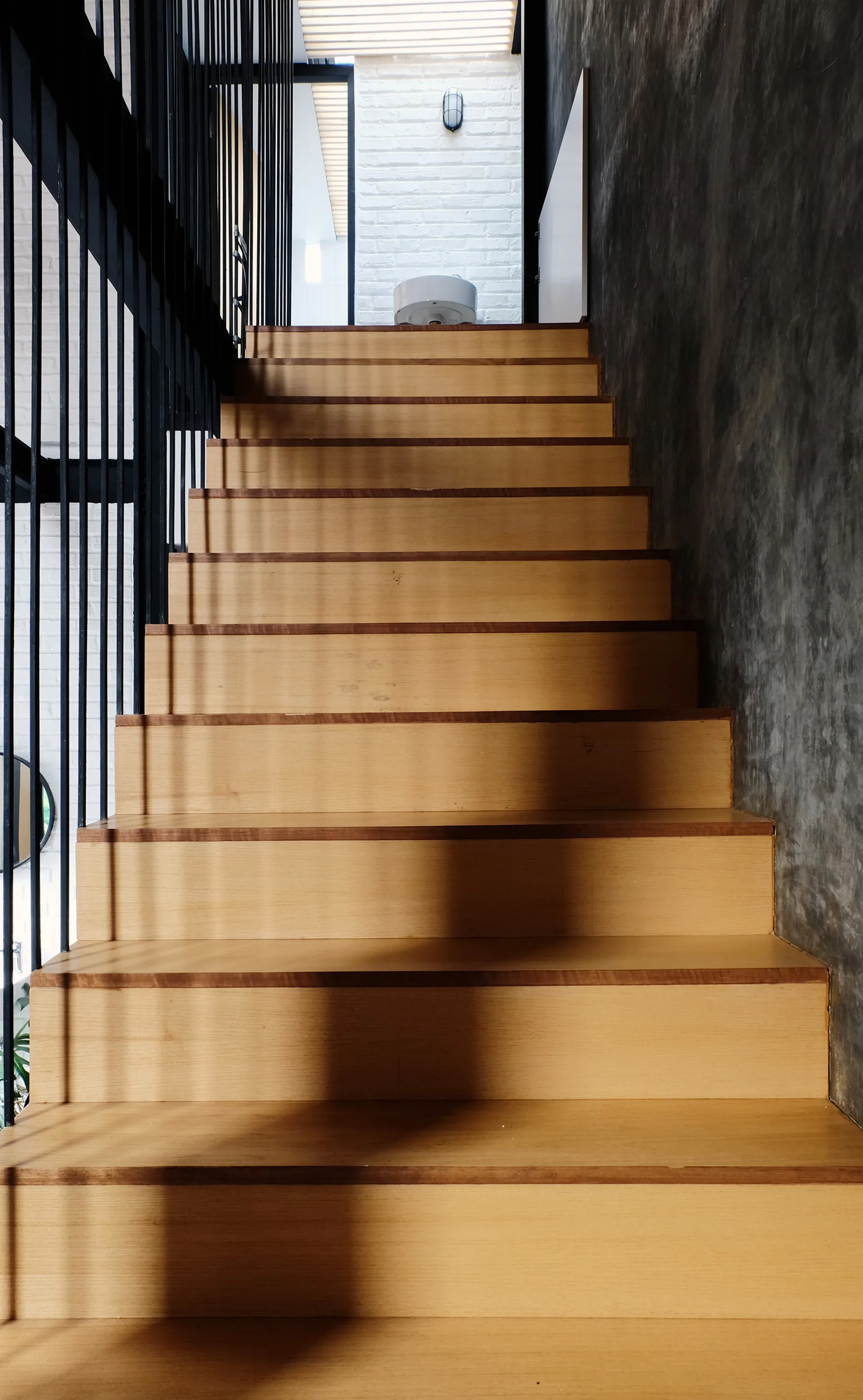 An eye-level view looking up a wooden staircase with a black vertical bar railing and a textured dark concrete wall.