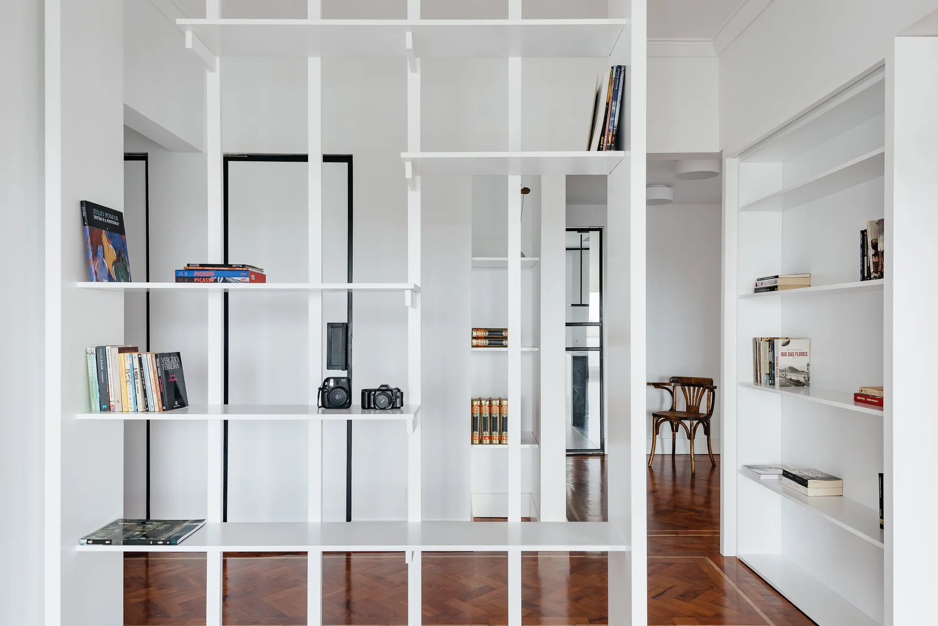 A large white open shelving unit acts as a room divider on dark wood herringbone parquet flooring, with a wooden chair visible in the background.