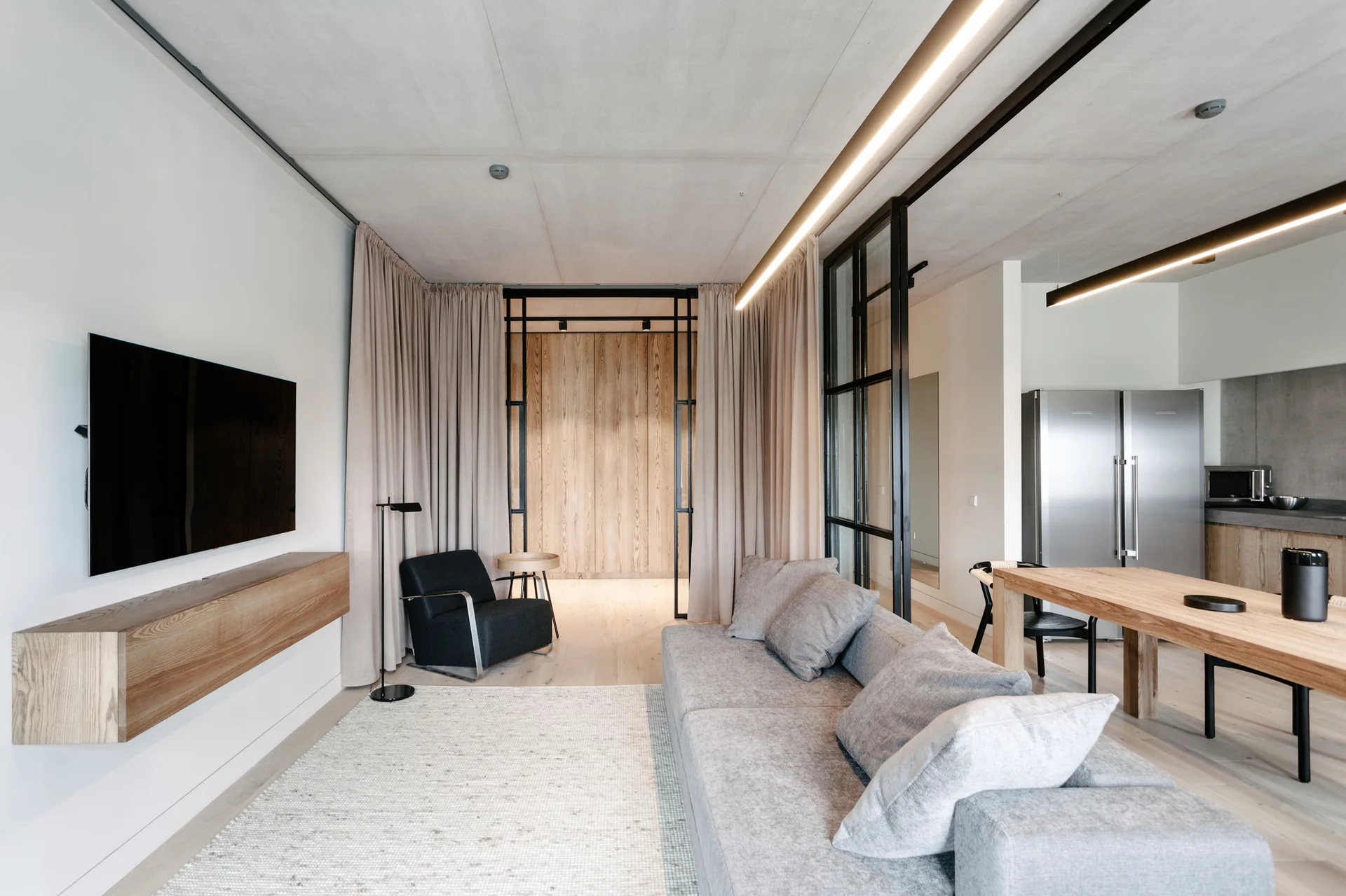 An open-concept living room features a grey sectional sofa, a floating wooden console, an industrial glass partition, and beige floor-to-ceiling curtains.
