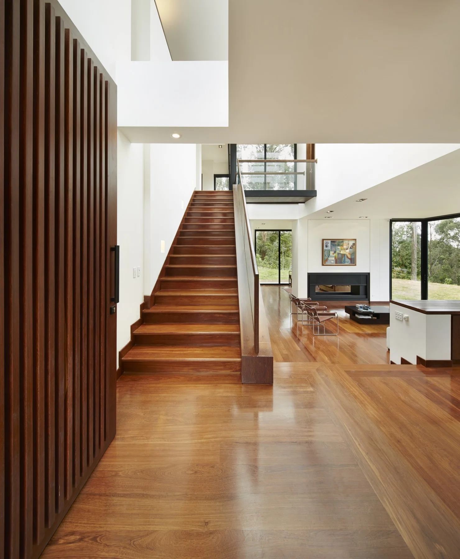 A contemporary entryway featuring a slatted wood door, polished hardwood floors, and a central staircase with a glass railing leading up.