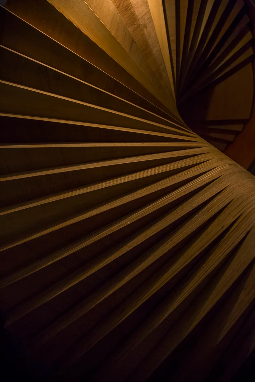 A close-up view of a helical spiral staircase made of stacked wooden steps with warm directional lighting casting shadows on the treads.